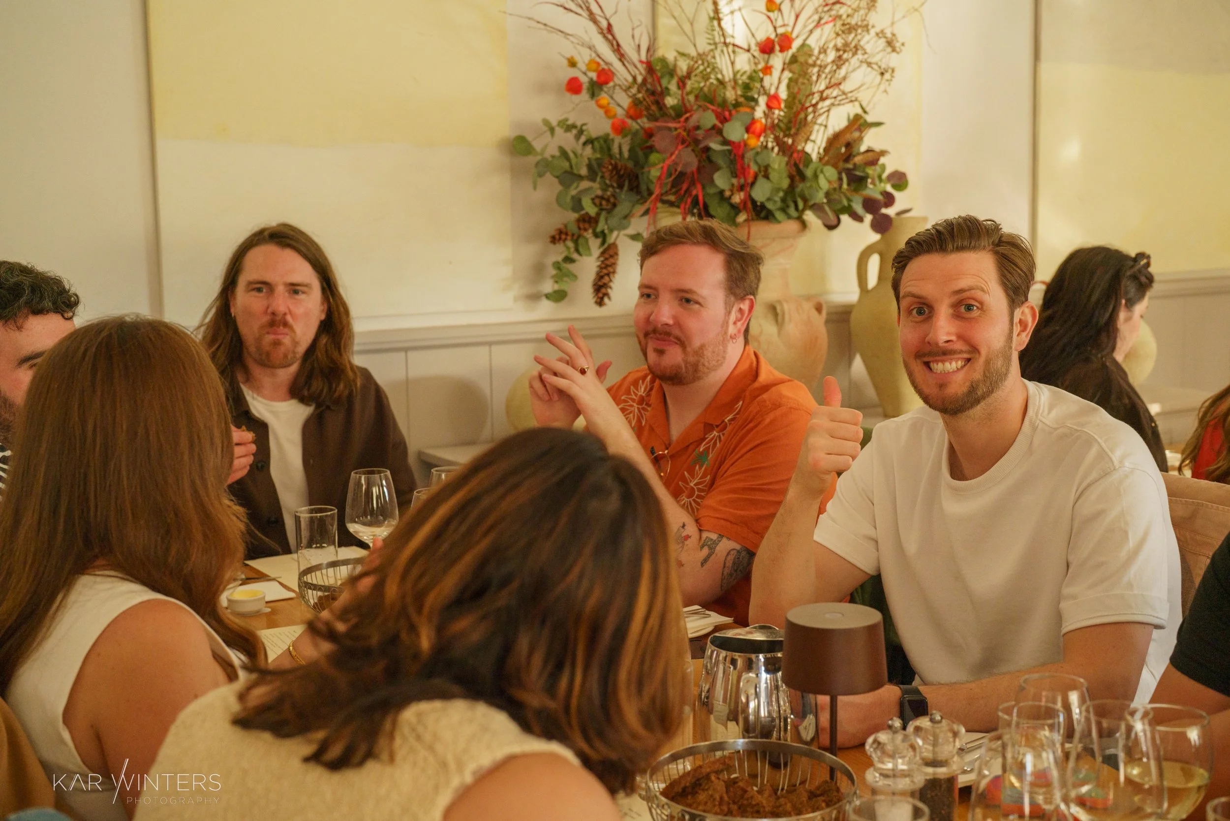Group of people sitting at a dinner table in a restaurant, smiling and talking. The table has glasses, a basket, and plates, with a decorative floral arrangement in the background.