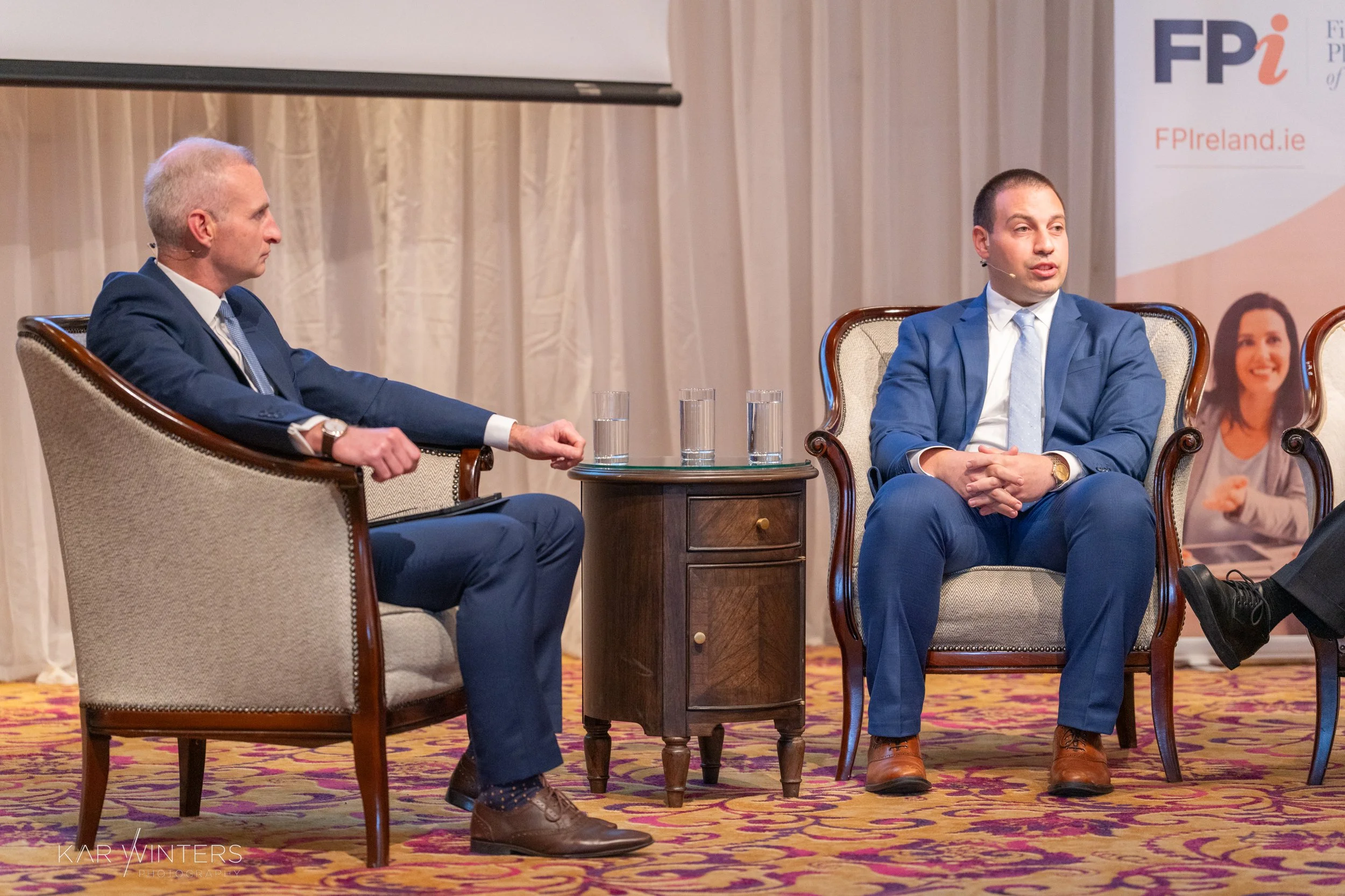 Two men in suits sitting on armchairs during a panel discussion, with three glasses of water on a small table between them, in a conference room with beige curtains and a banner in the background.