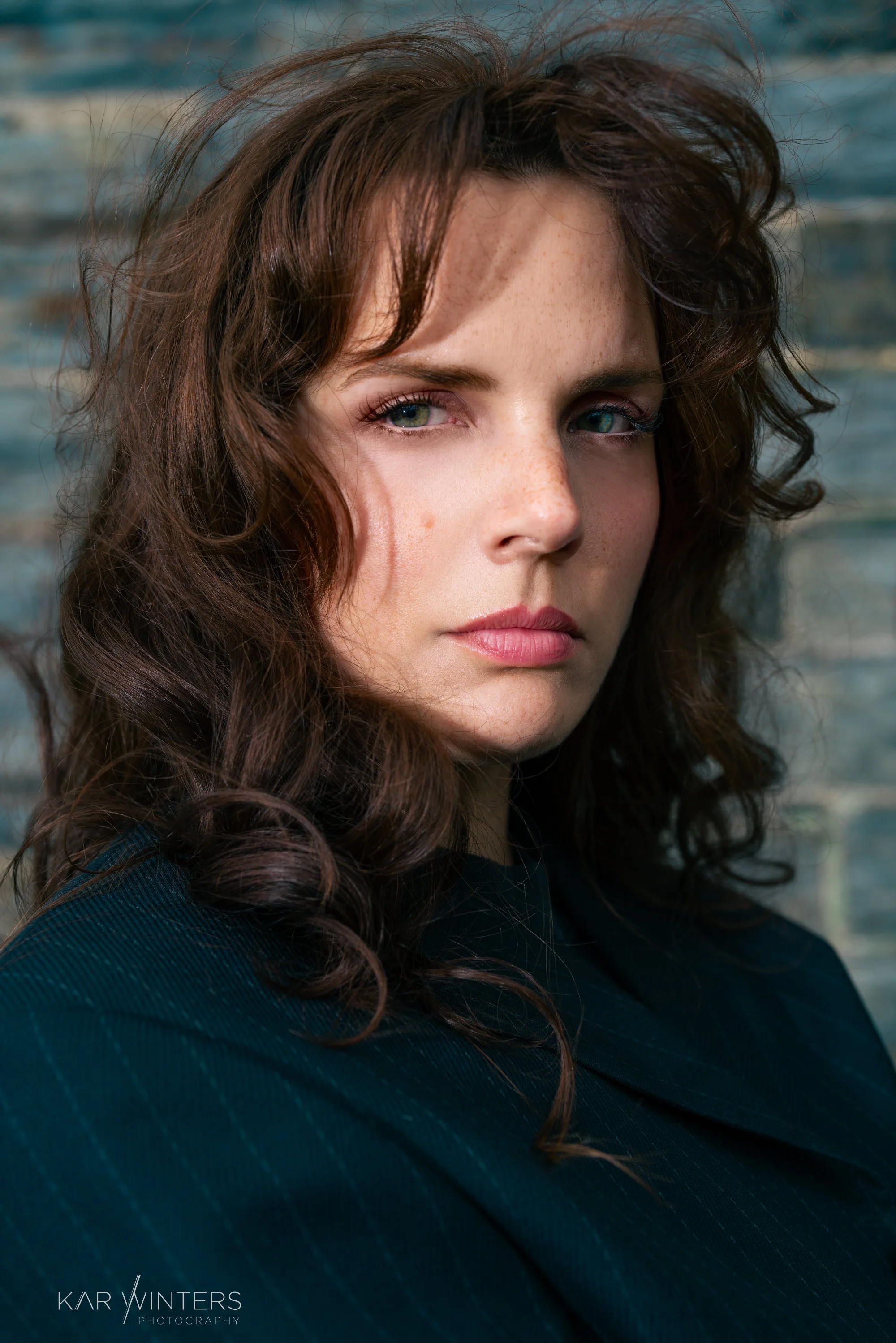 Close-up portrait of a woman with curly brown hair, blue eyes, and freckles, looking directly at the camera against a blue and gray outdoor background.