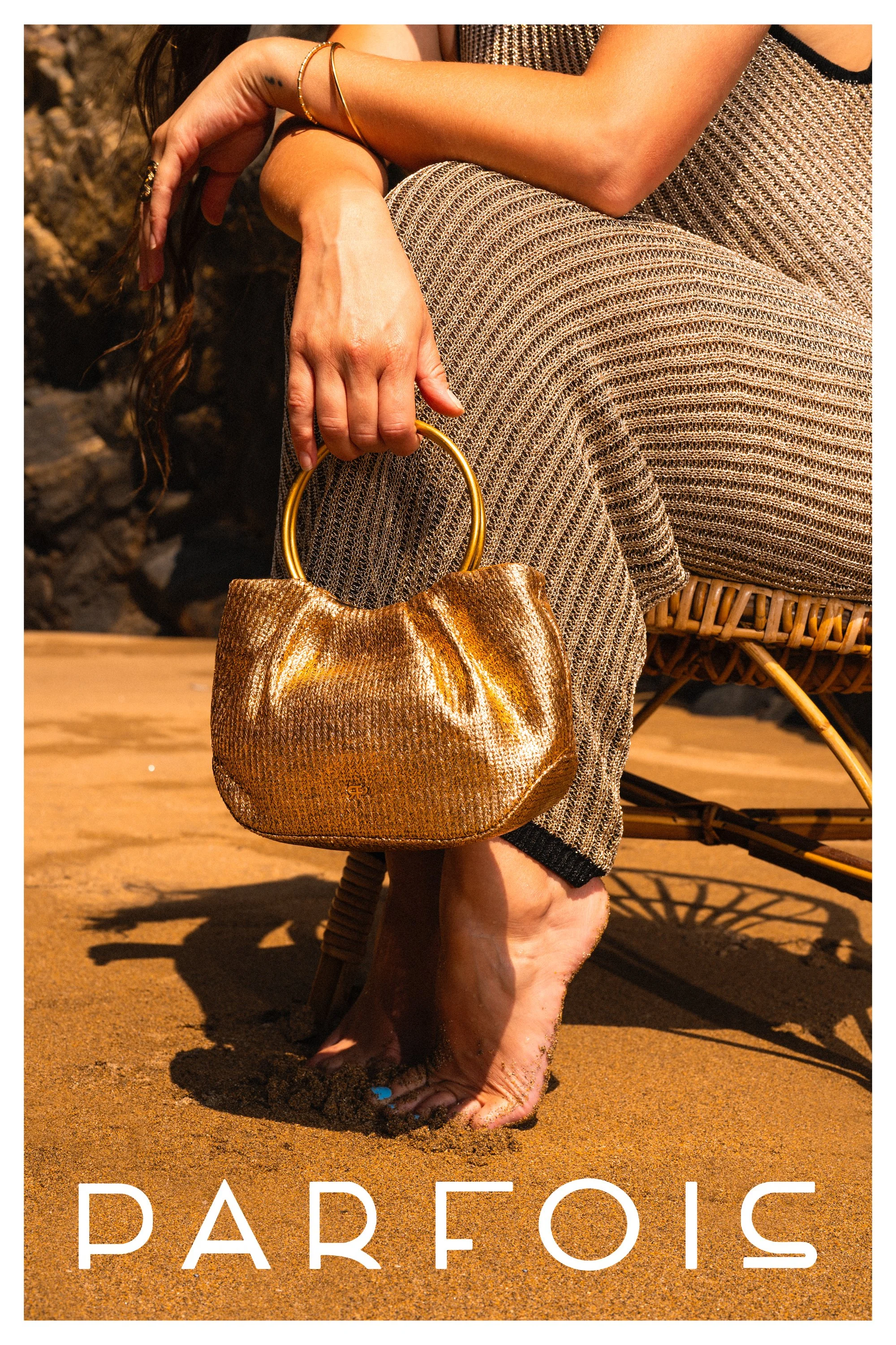 A woman is sitting barefoot on a wooden chair, holding a shiny, gold purse with a circular handle. She is wearing a textured, patterned dress and some gold jewelry, and is sitting on sand with rocks in the background. The word 'PARFOIS' is text at th