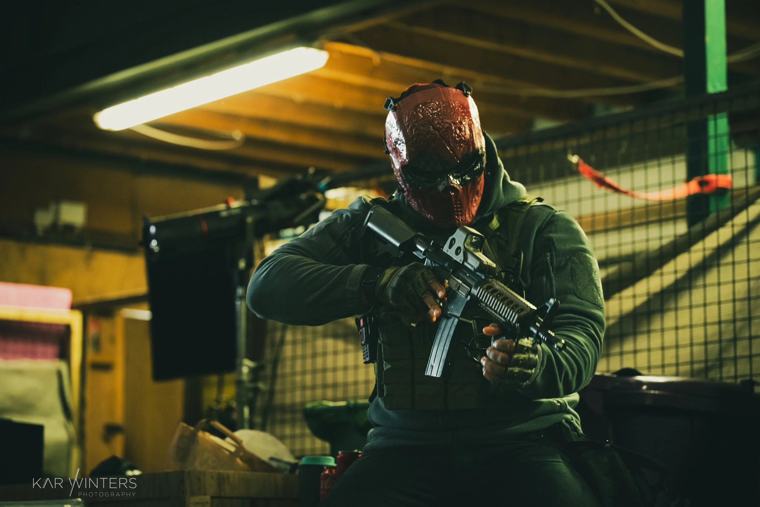Person wearing a red mask and tactical gear holding a rifle inside a room with a grid wall and fluorescent lighting.