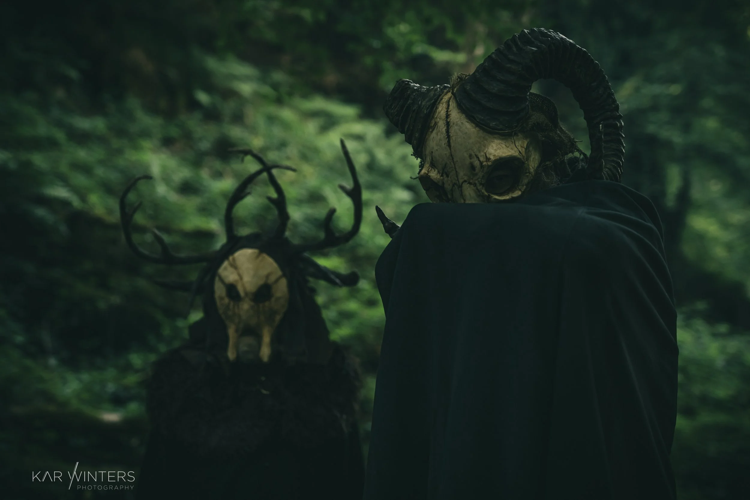 Two figures wearing animal skull masks with horns, one with a person's body and the other with a large antlered skull, in a dark green forest setting.