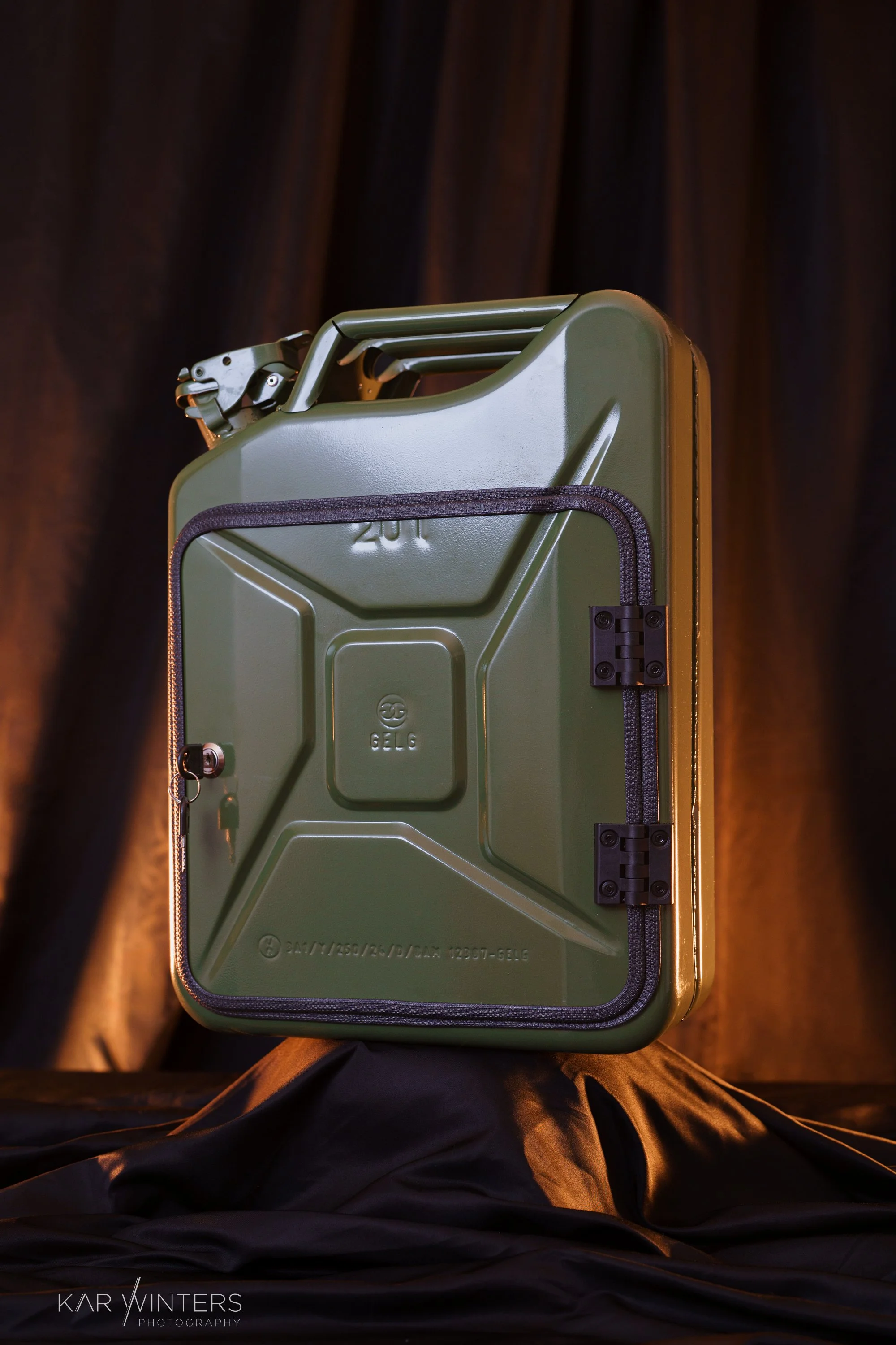A green military-style metal case with black trim, placed on a black cloth with a dark brown curtain background.