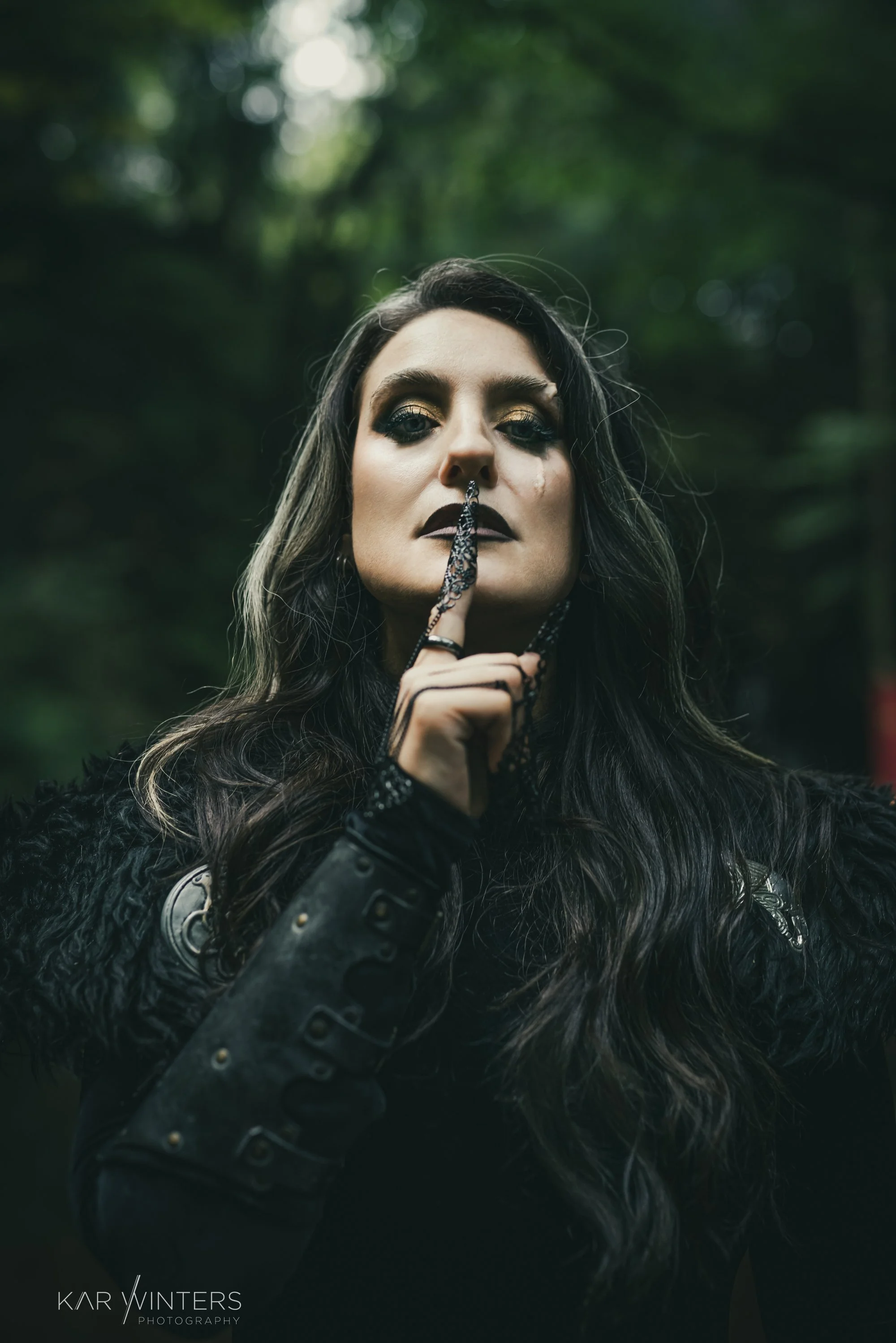 A woman with long dark hair and dark makeup, holding a chain with a lock to her lips, standing outdoors in a forest with a serious expression.