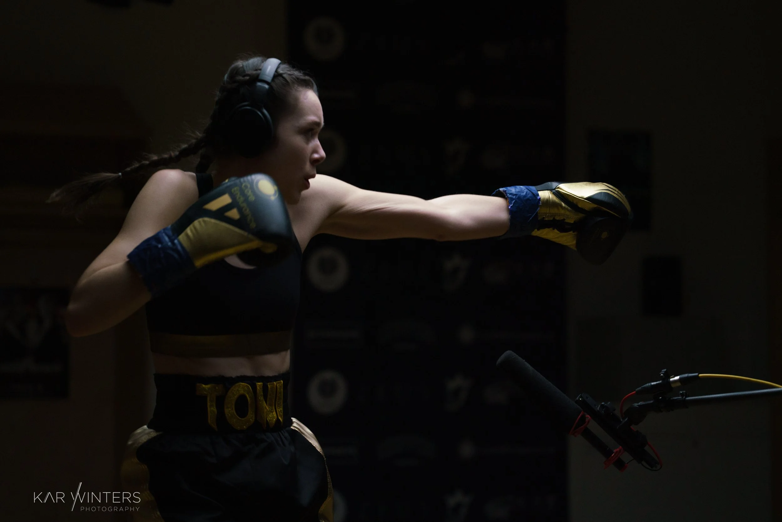 A female boxer practicing punches in a training studio, wearing boxing gloves, headphones, and boxing shorts labeled 'TOW', with her left arm extended in a punch.