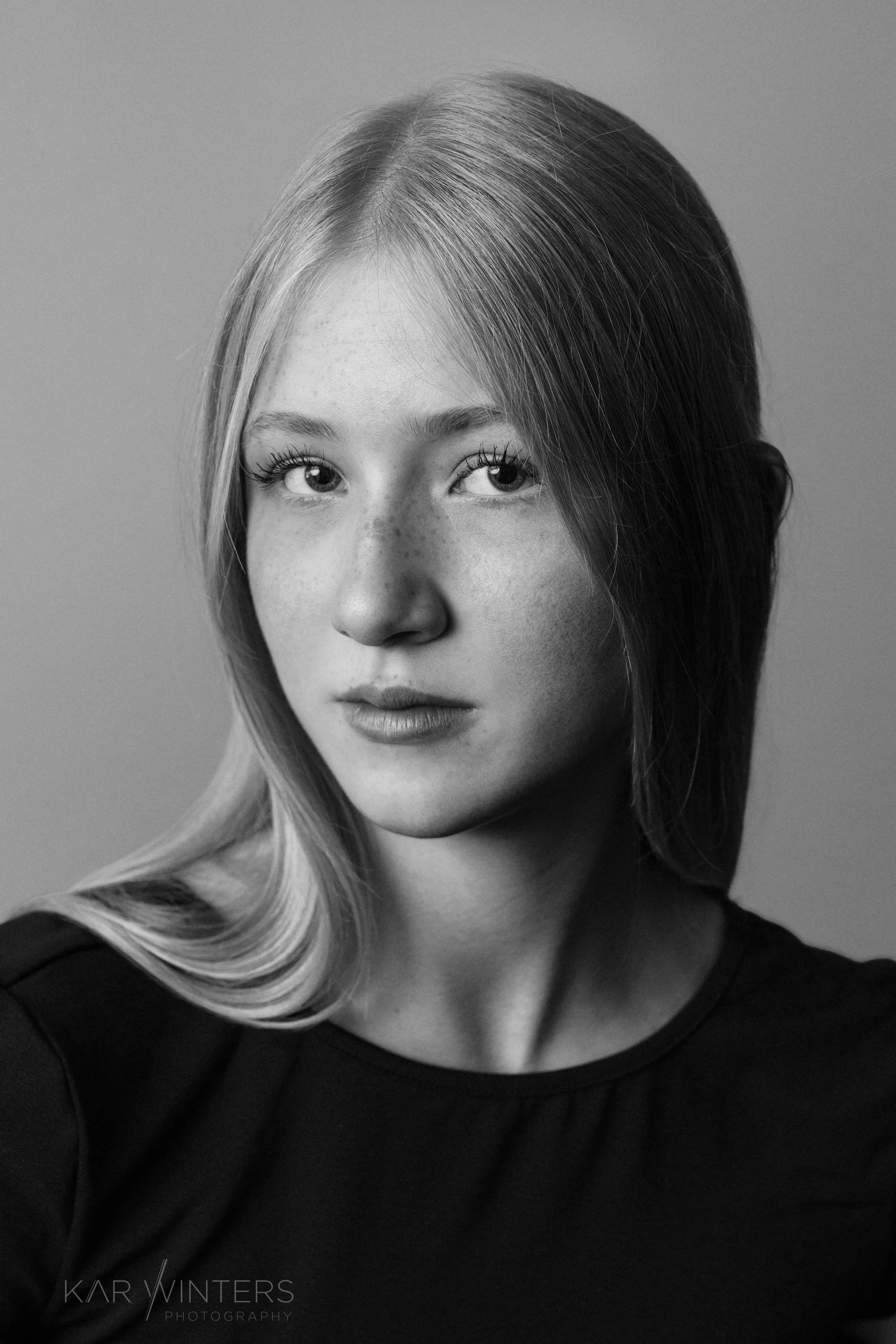 Black and white portrait of a young woman with light skin, freckles, and long wavy hair, wearing a dark top, looking directly at the camera.