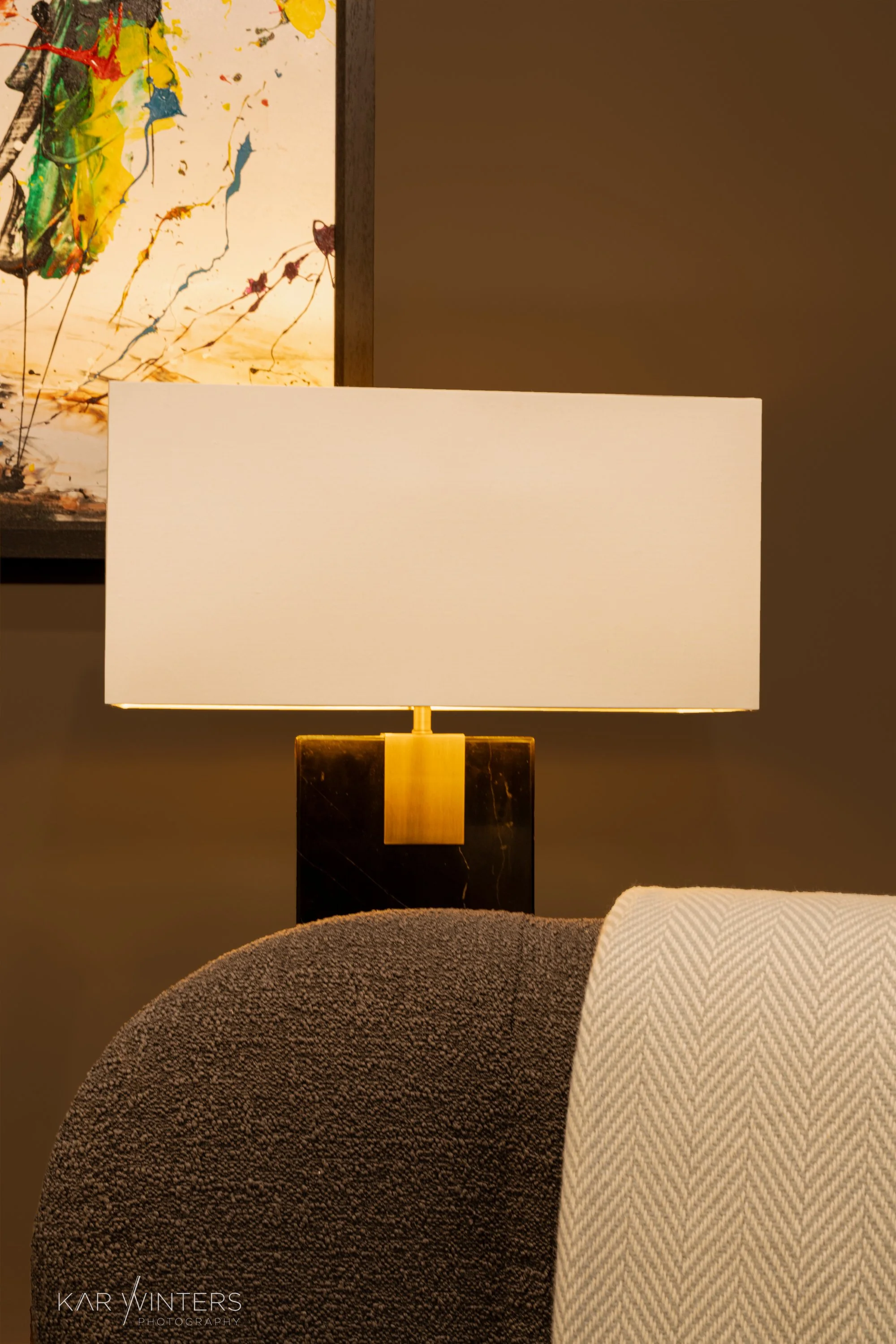 A modern table lamp with a rectangular ivory shade and black marble base with gold accent, placed on a table with curved brown and beige upholstered chairs. An abstract colorful painting with splashes of yellow, green, and blue is on the wall behind 