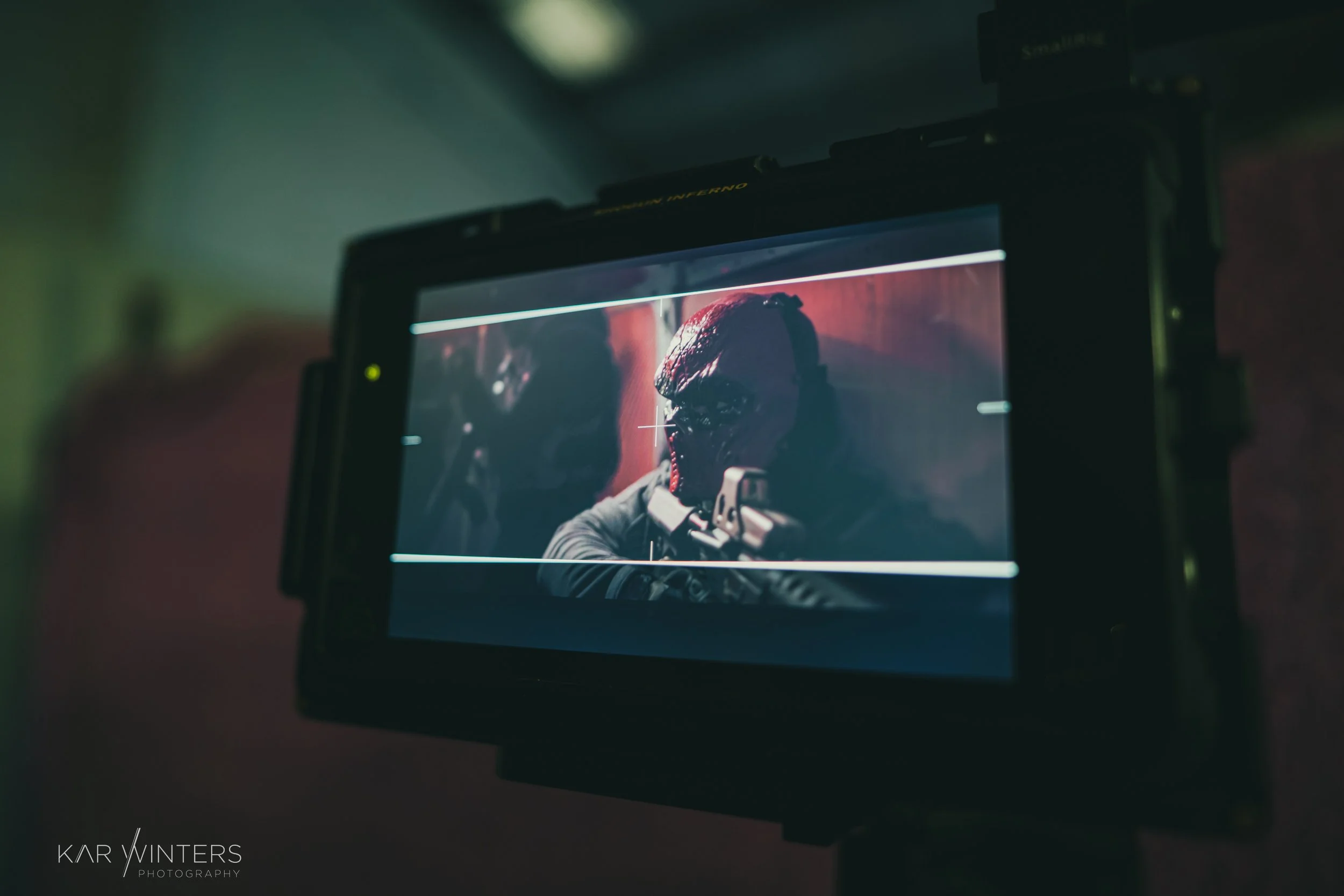 A scene from a film or photoshoot showing a person in a mask aiming a gun, photographed through a camera monitor.