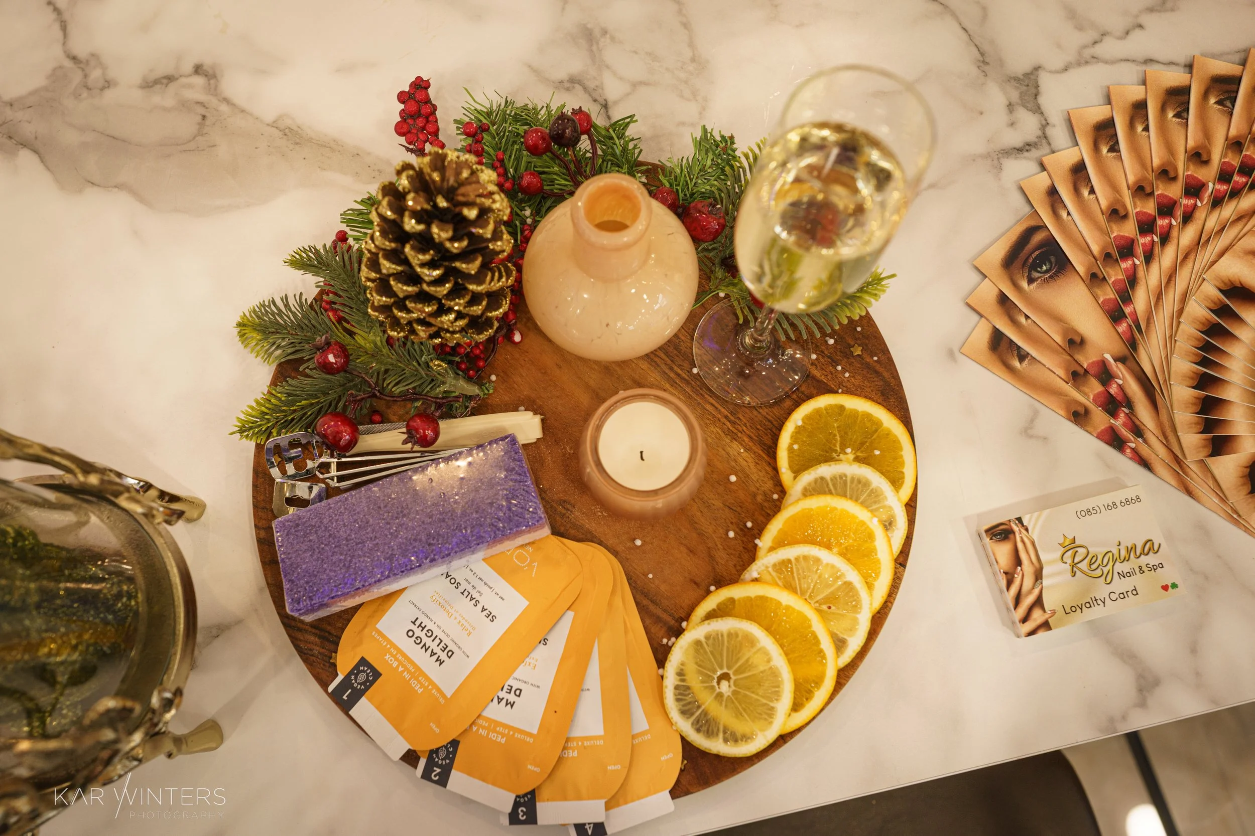 Decorative table display with a pinecone, red berries, greenery, a candle, slices of lemon and orange, a glass of champagne, business cards, and promotional flyers for a nail and spa salon.