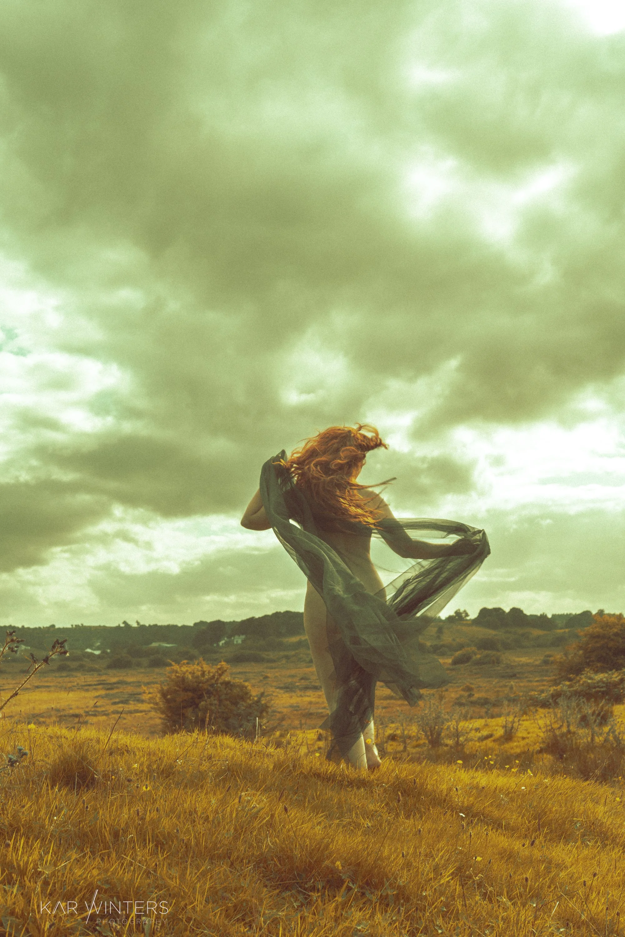 A woman with red hair standing in a grassy field under cloudy skies, holding a transparent black fabric around her.