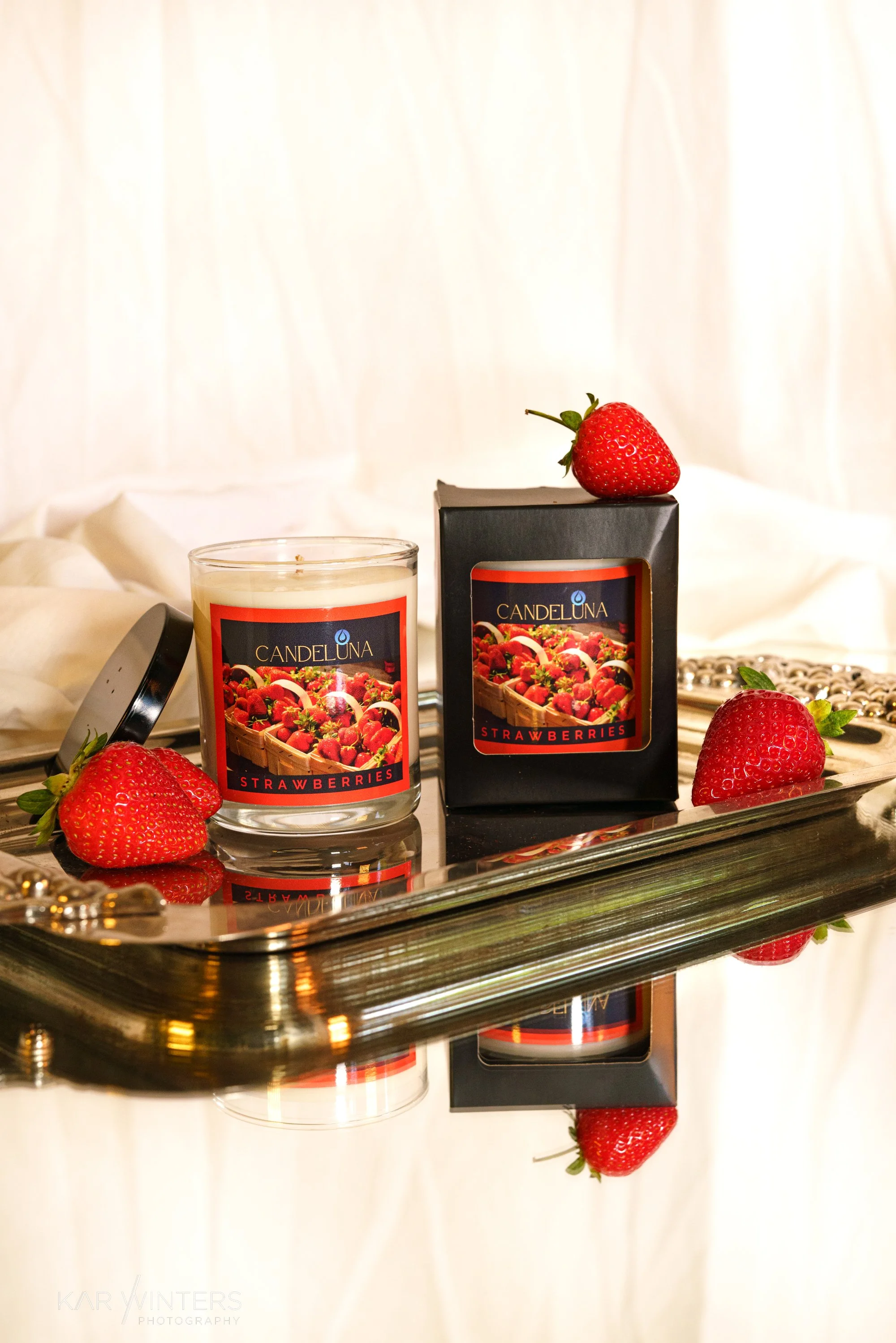 A scented candle labeled 'Candeluna' with a strawberry on top of its black box, along with three fresh strawberries, all placed on a reflective silver tray.