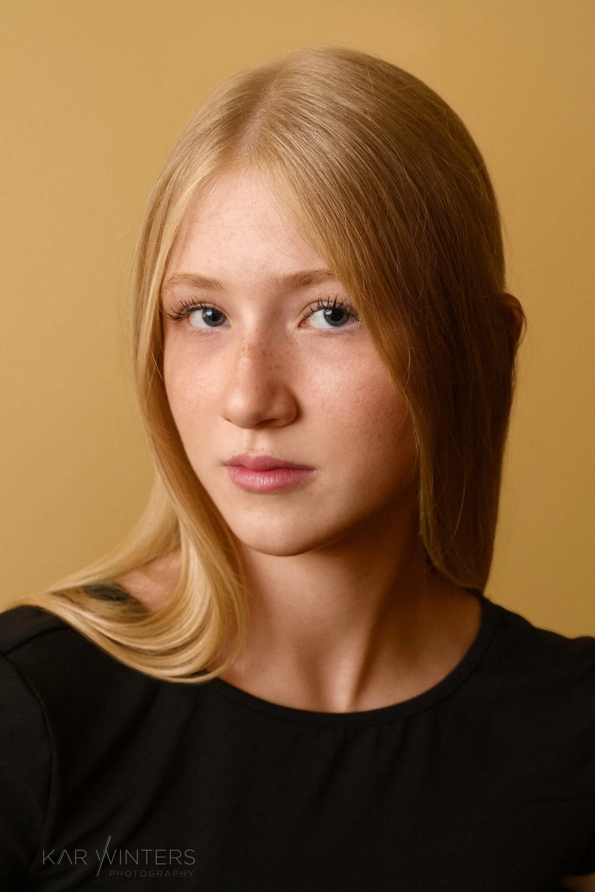 Close-up portrait of a young woman with red hair and blue eyes, wearing a black top, against a beige background.