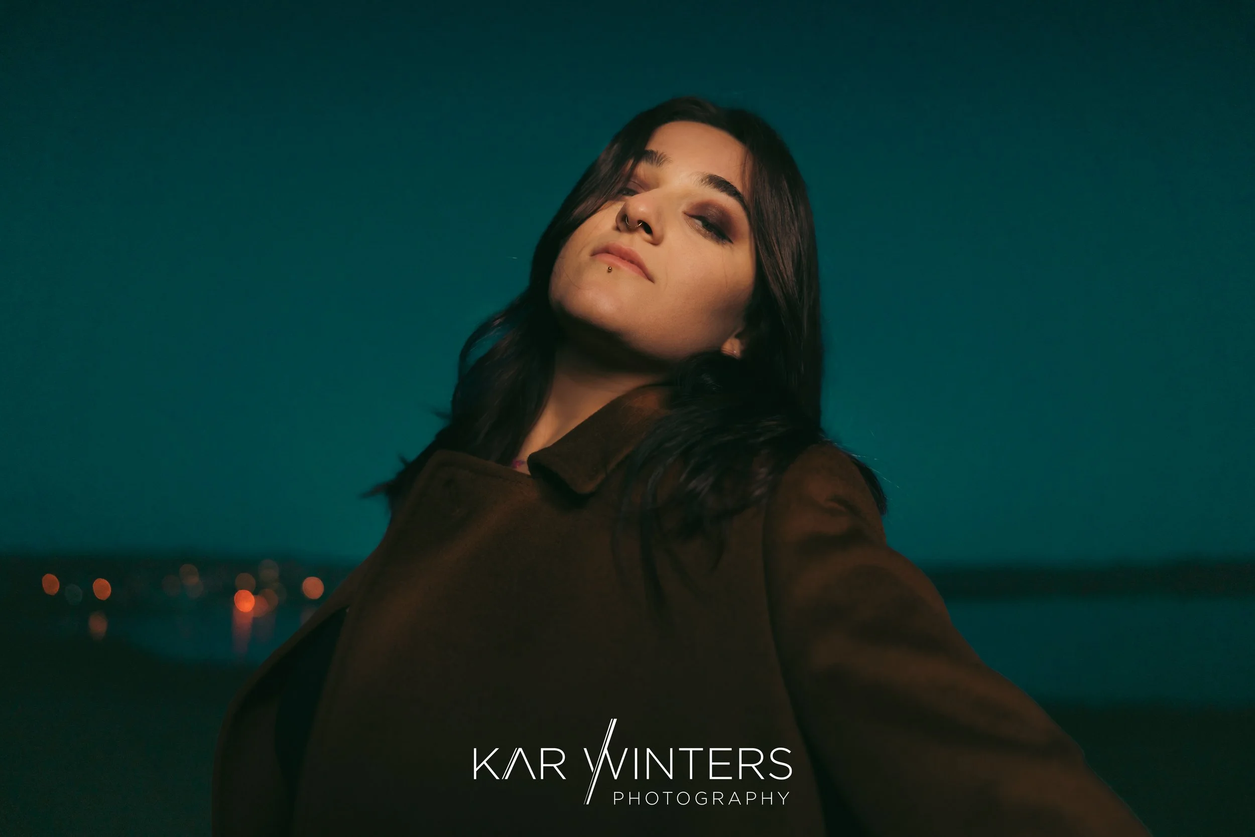 A woman with dark hair, makeup, and piercings taking a selfie outdoors at night, with a body of water and city lights in the background.