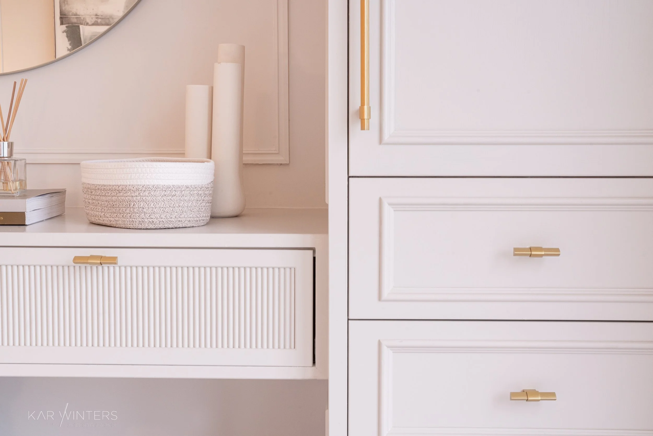 Close-up of a white dresser with gold handles, decorated with a round white textured basket, two tall white vases, and a glass jar with reed sticks, and a book. Part of a mirror and wall trim are visible in the background.