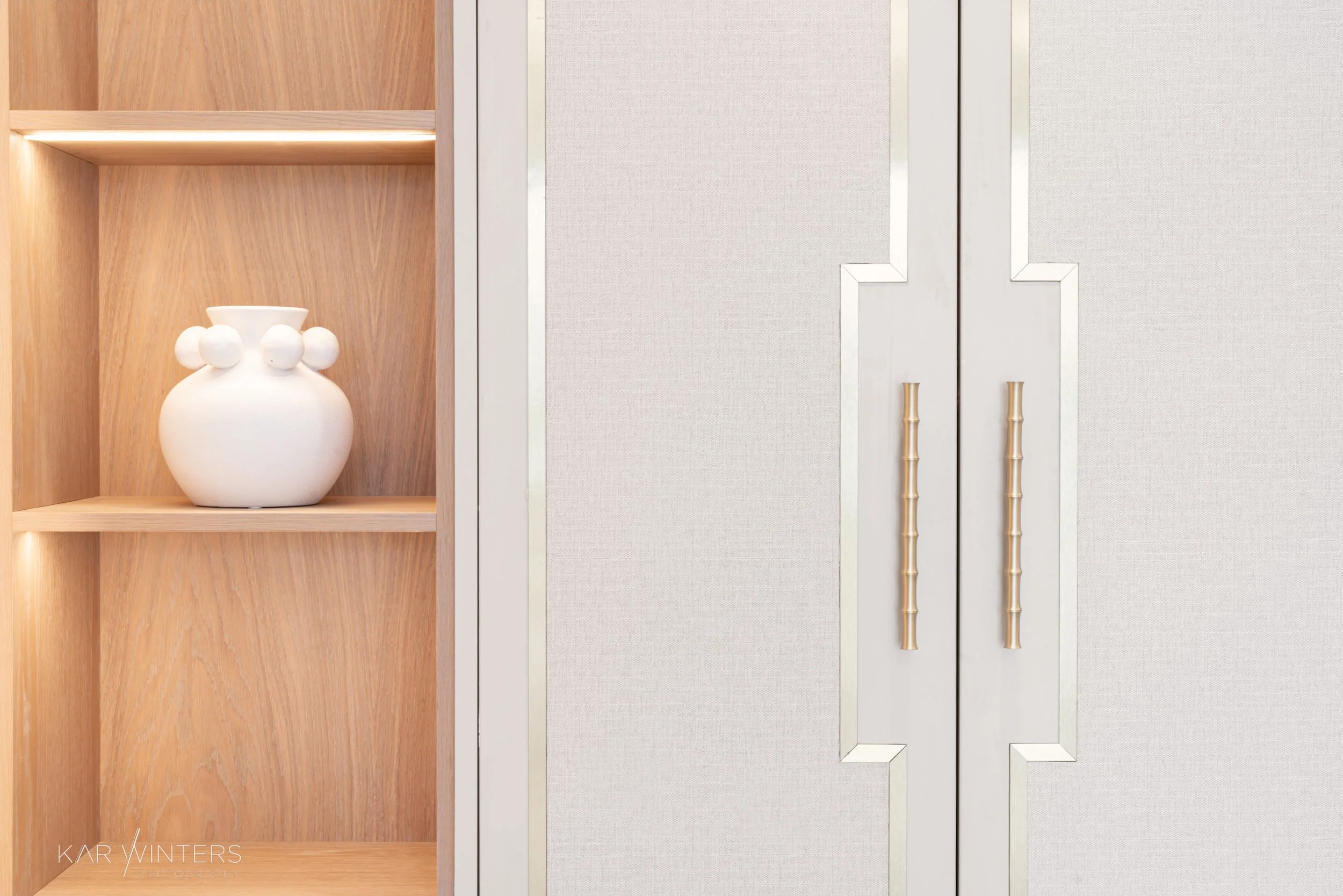 A white ceramic vase with a rounded body and spherical handles on two sides sitting on a wooden shelf, next to closed cabinet doors with gold bamboo-style handles.