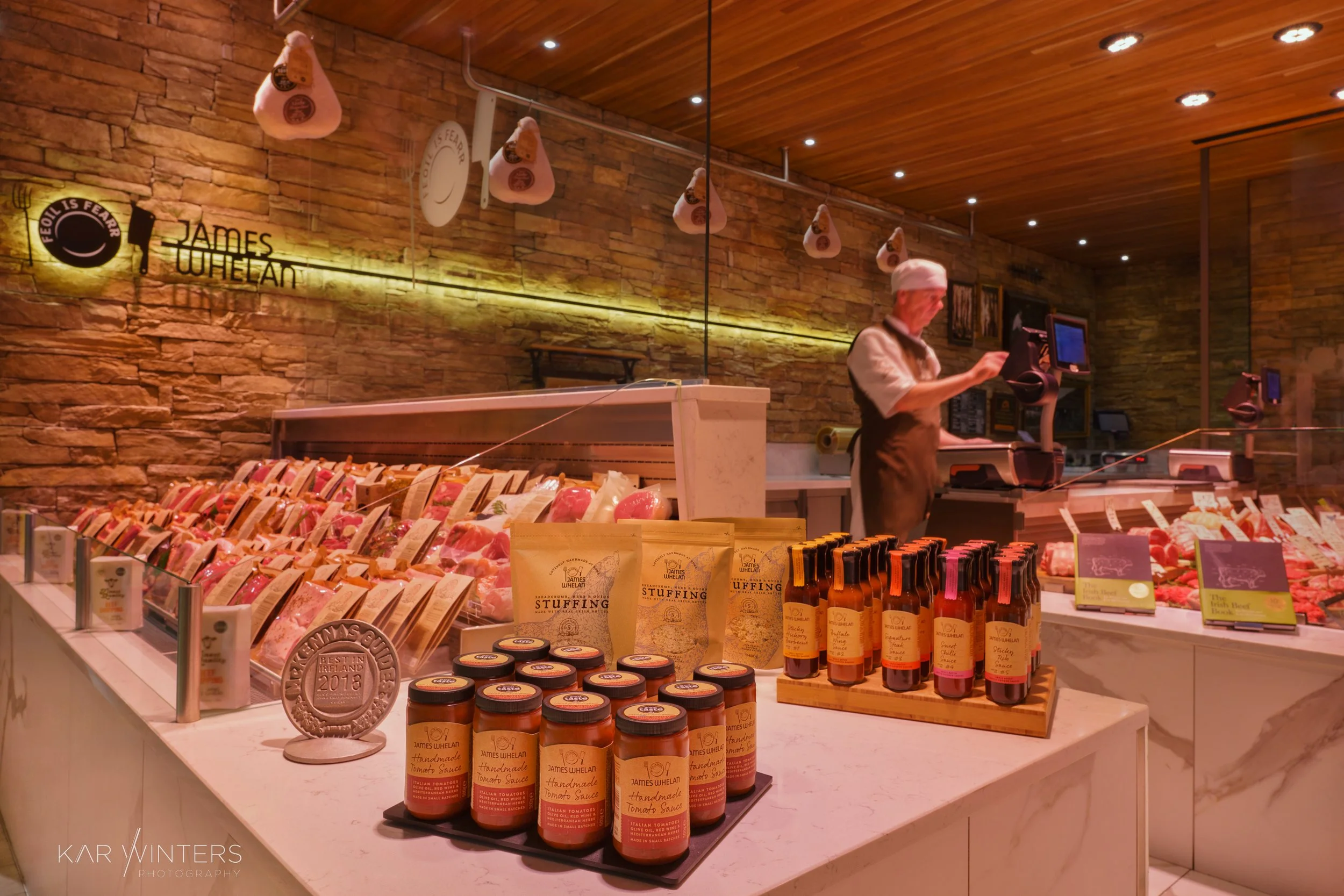 A butcher shop with various cuts of meat displayed behind a marble counter, a staff member in apron and hat working at the cash register, and shelves with condiments and sauces, all inside a warmly lit room with stone walls and wooden ceiling.