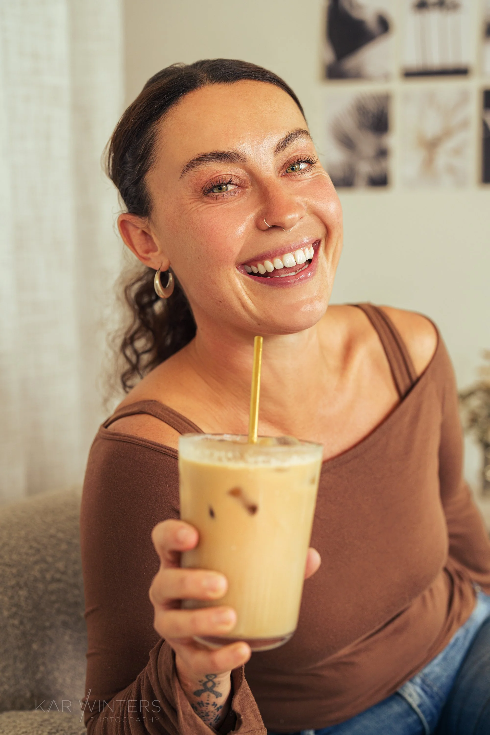 A smiling woman with dark hair, wearing hoop earrings and a septum piercing, holding a glass of iced coffee with a straw. She is wearing a brown top and has a tattoo on her arm.
