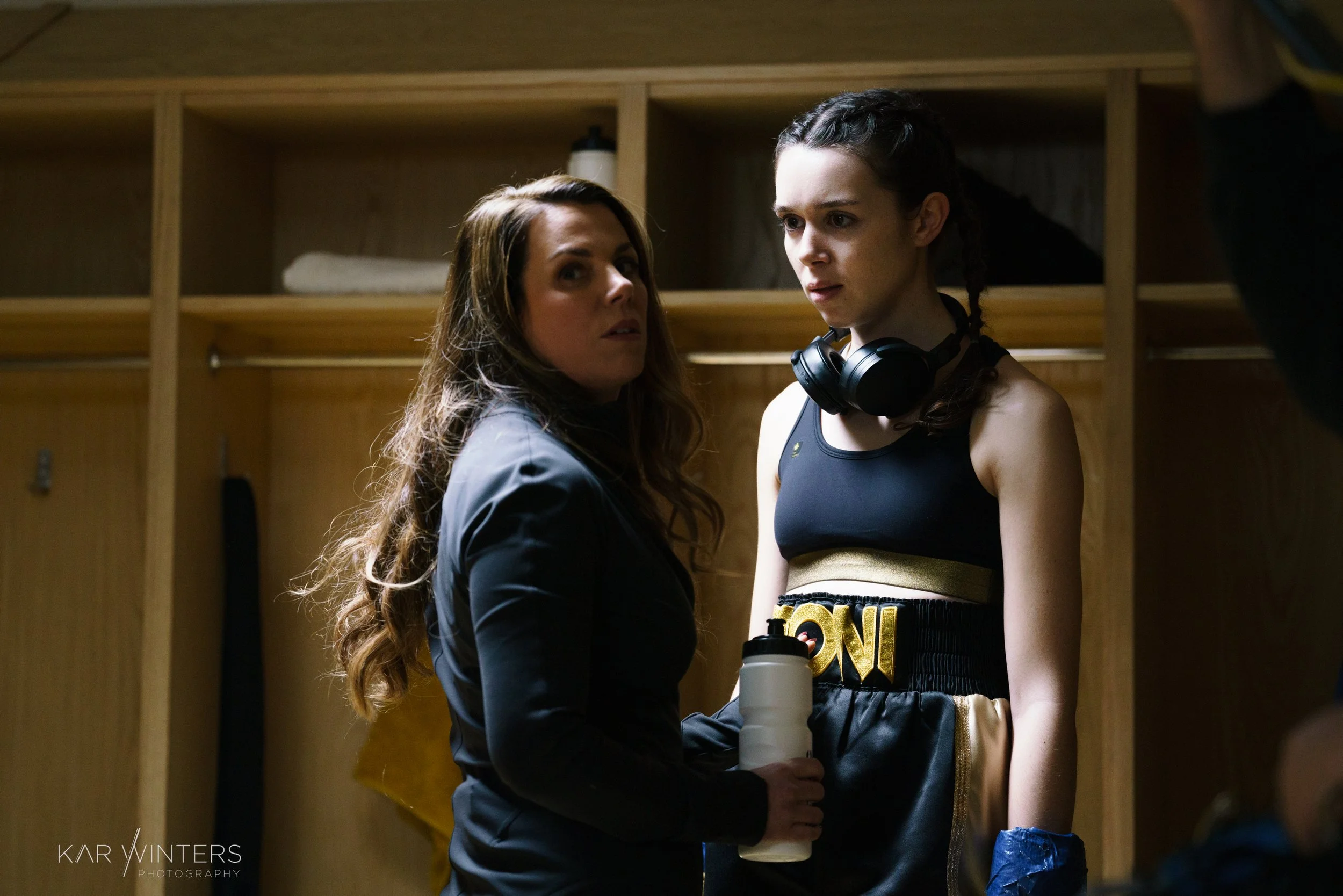 Two women in a gym locker room, one dressed in boxing attire with headphones around her neck and a water bottle in hand, the other dressed in casual clothing, appear to be talking.
