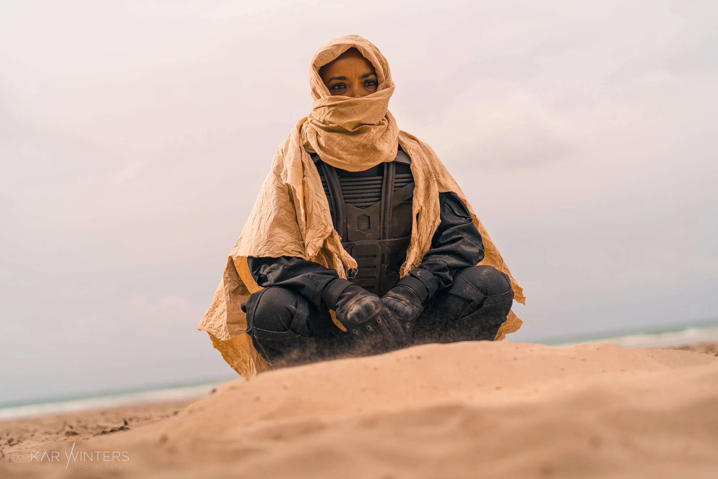 A person dressed in tactical gear and a tan hooded cloak, sitting on a sandy beach with a cloudy sky in the background.