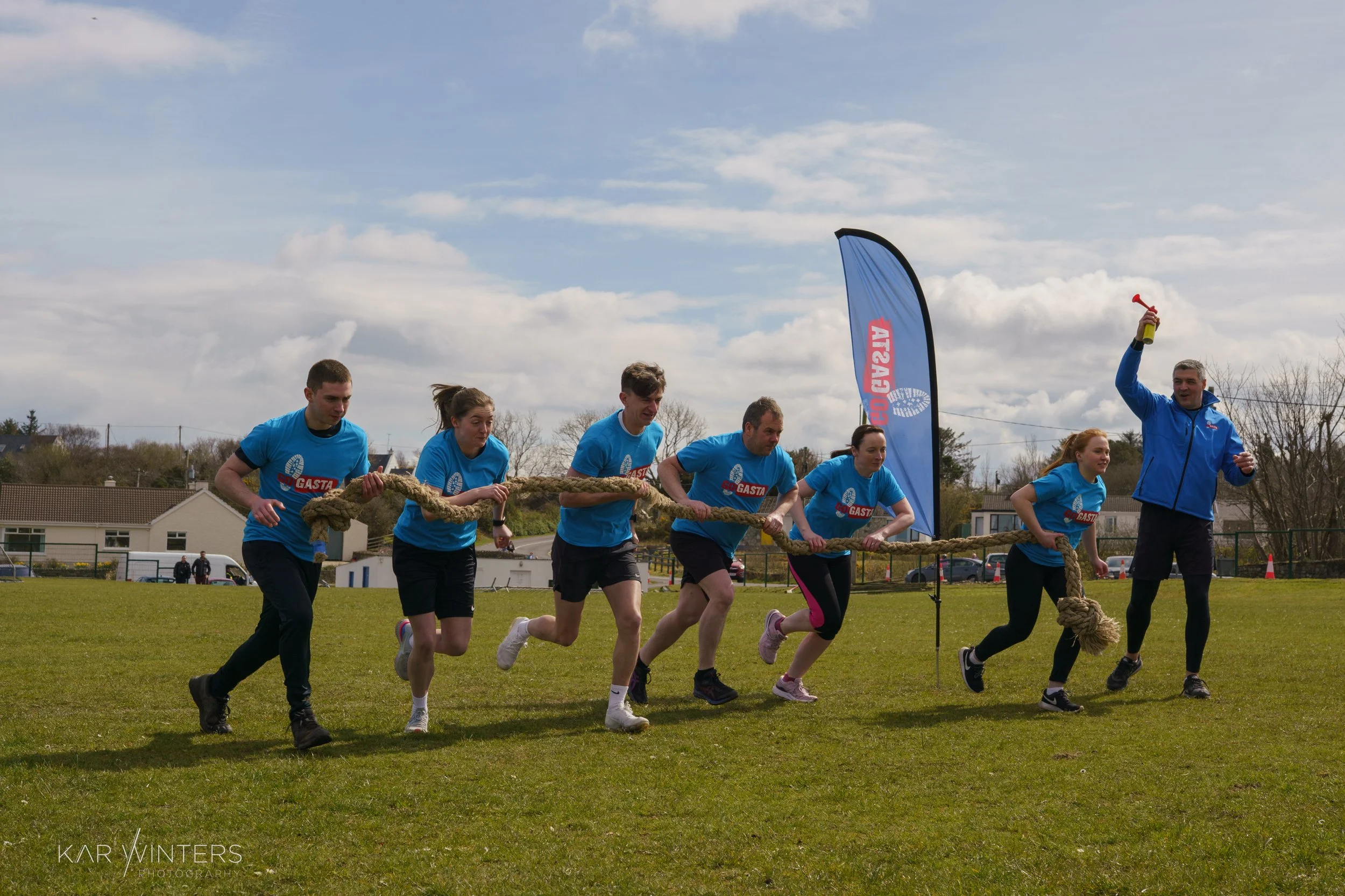 Group of six people participating in a tug-of-war contest outdoors on a grassy field, with a man holding a yellow horn in one hand and a flag in the background.