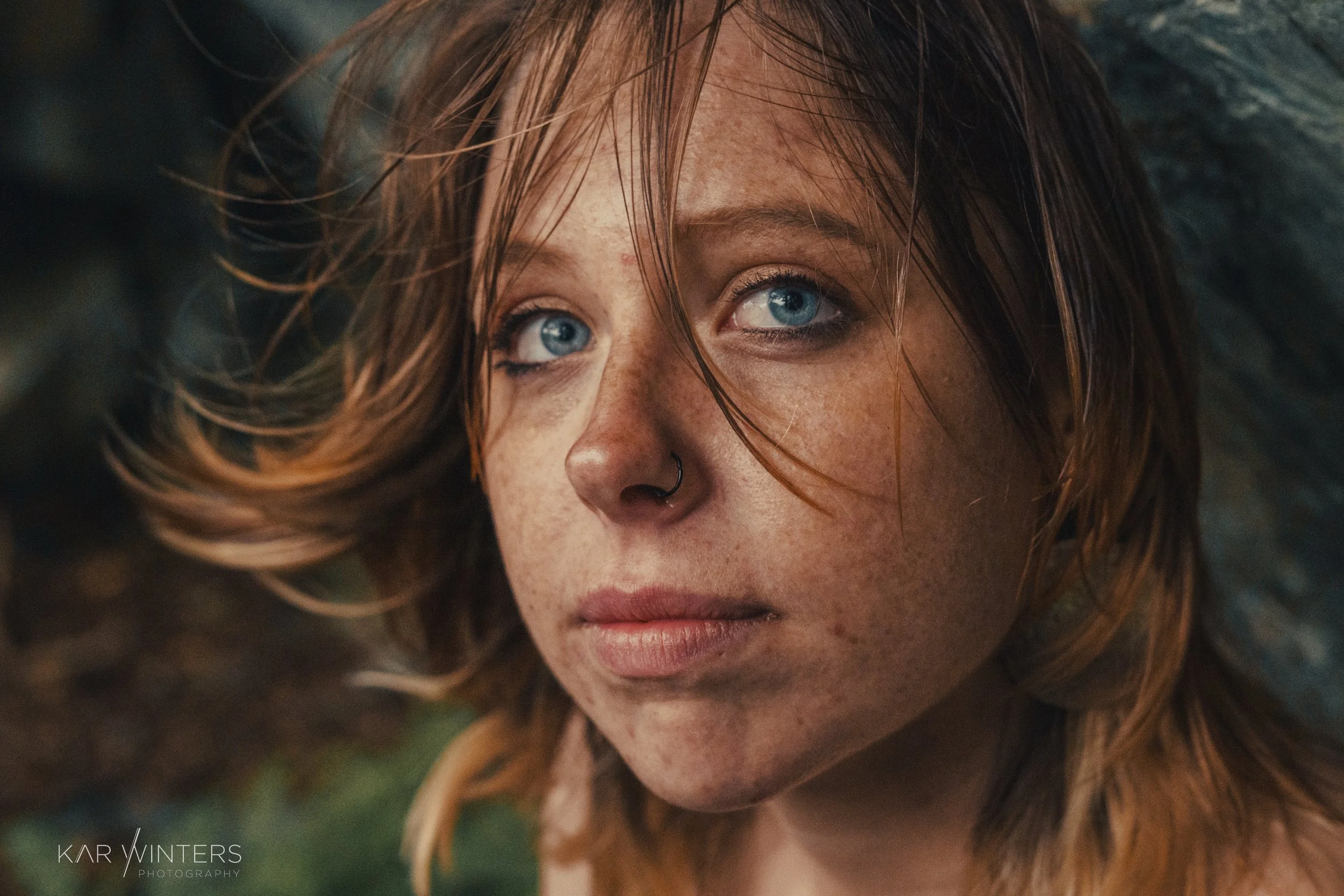 A close-up portrait of a young woman with red hair, blue eyes, freckles, and a septum piercing, looking intently at the camera with a natural outdoor background.