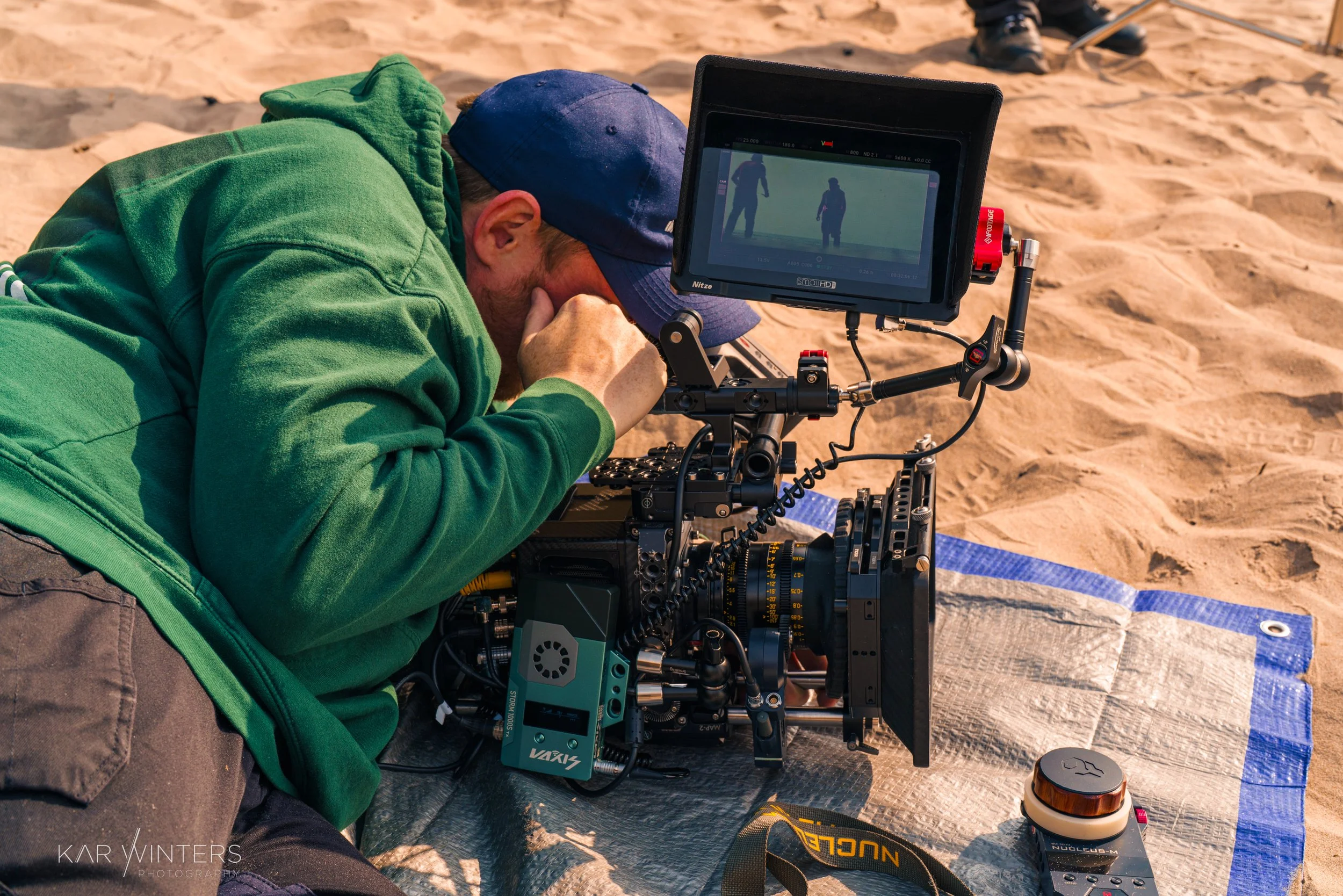 A film crew member lies on the sand, looking through the viewfinder of a professional camera mounted on a tripod, with a monitor attached. The scene appears to be on a sandy beach with some equipment nearby.