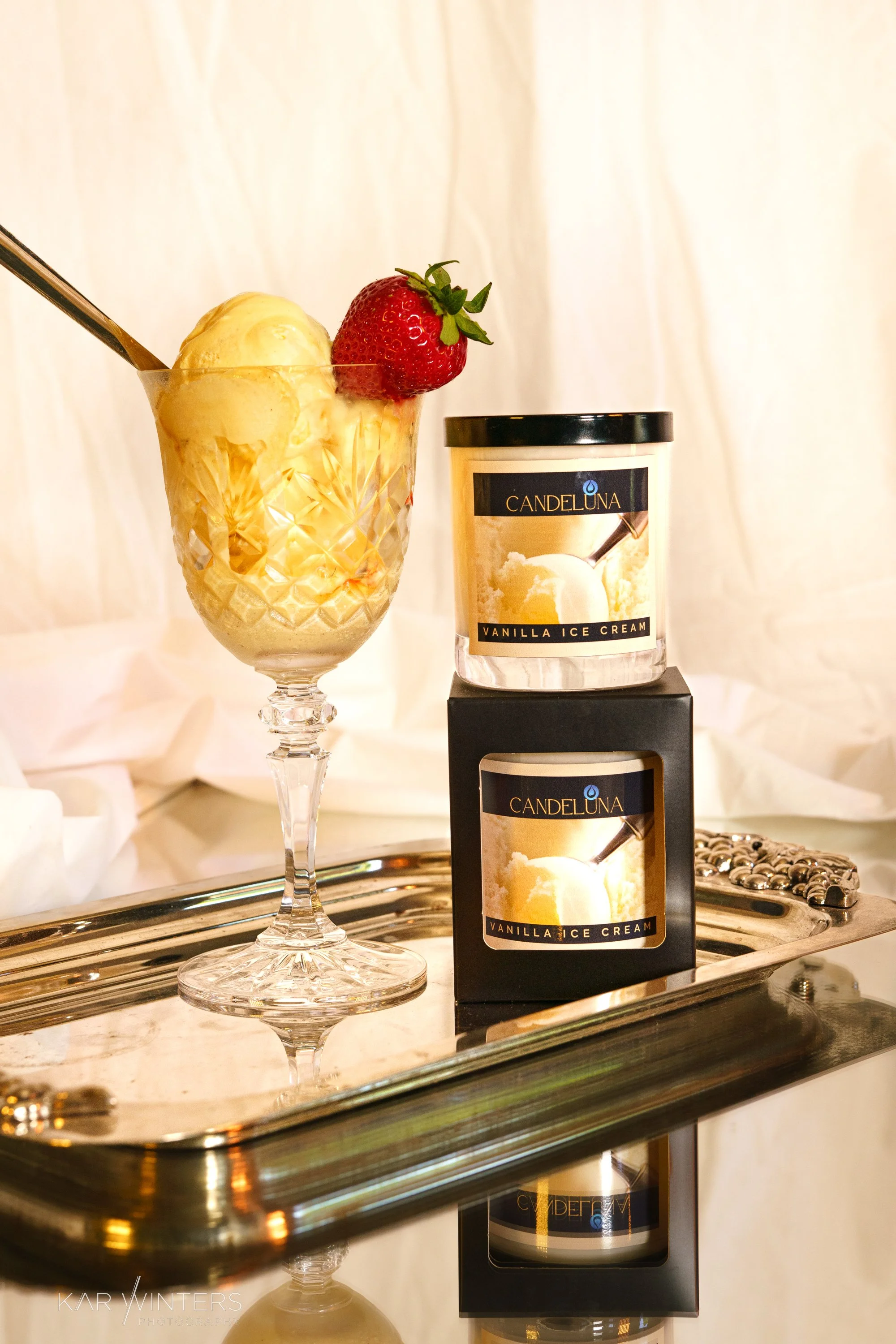 A glass of vanilla ice cream topped with a strawberry and a spoon, placed on a silver tray with two boxes of Candeluna vanilla ice cream behind it.