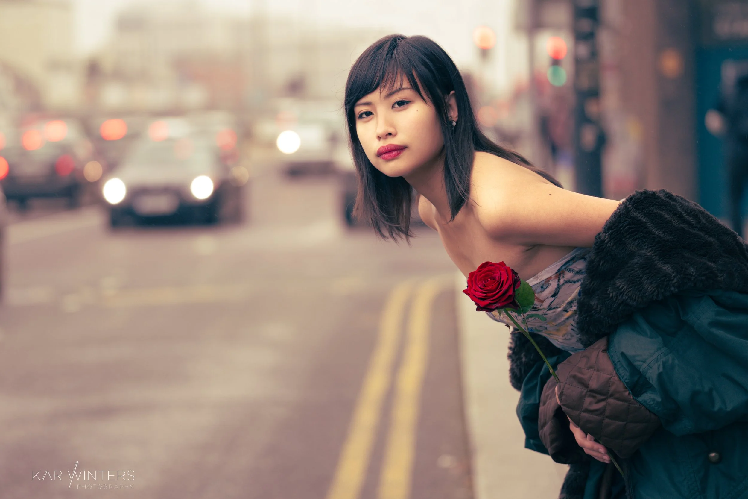 A woman with dark hair and a light makeup look, holding a red rose in front of her chest, leaning forward on a city street at dusk. She is wearing a patterned strapless top and a dark coat draped over her arm, with blurred cars and streetlights in th