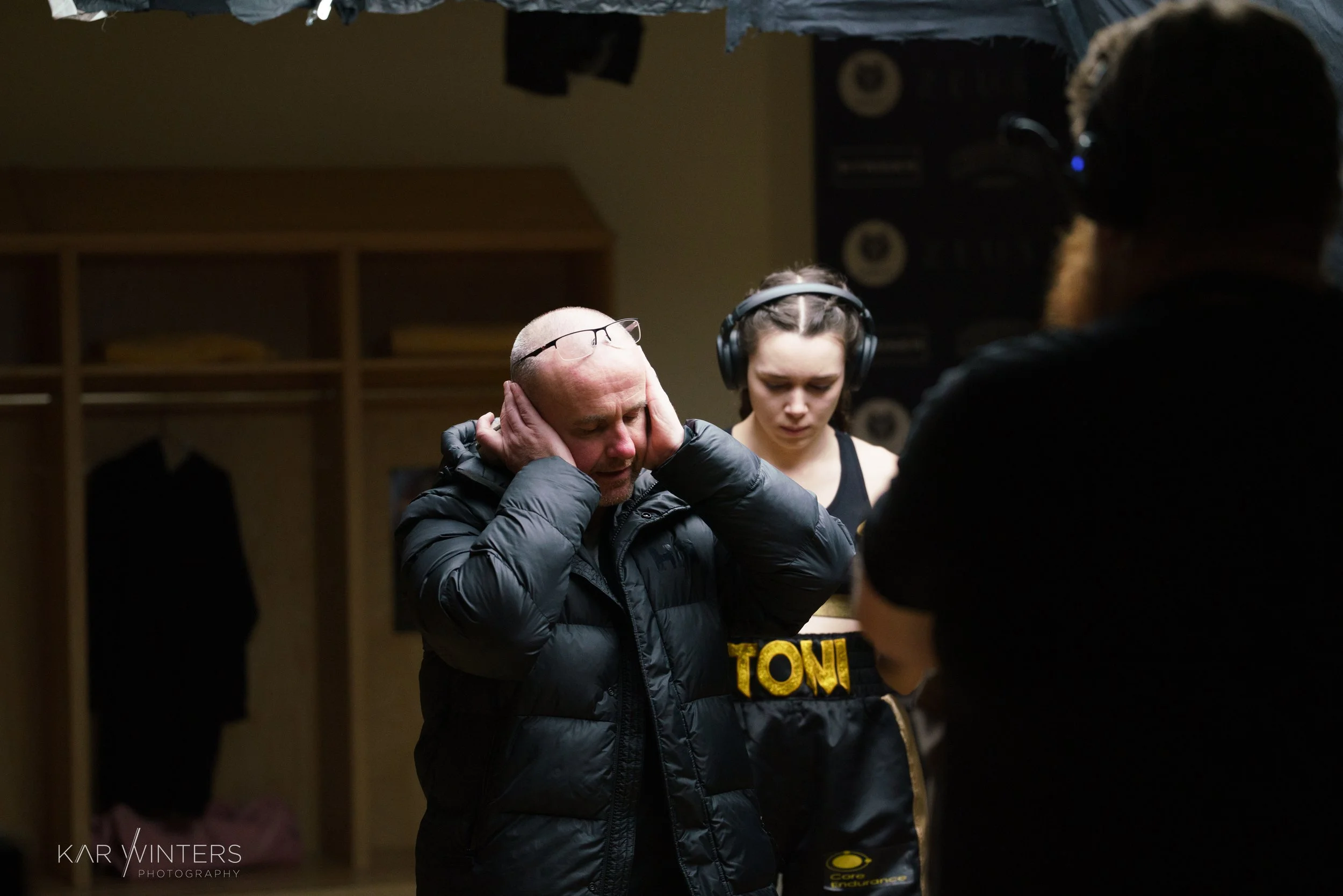 A man with glasses on his head and a woman with large headphones standing with two other individuals, appears to be in a gym or sports locker room, with one person in front and one on the right, all focused on the man.