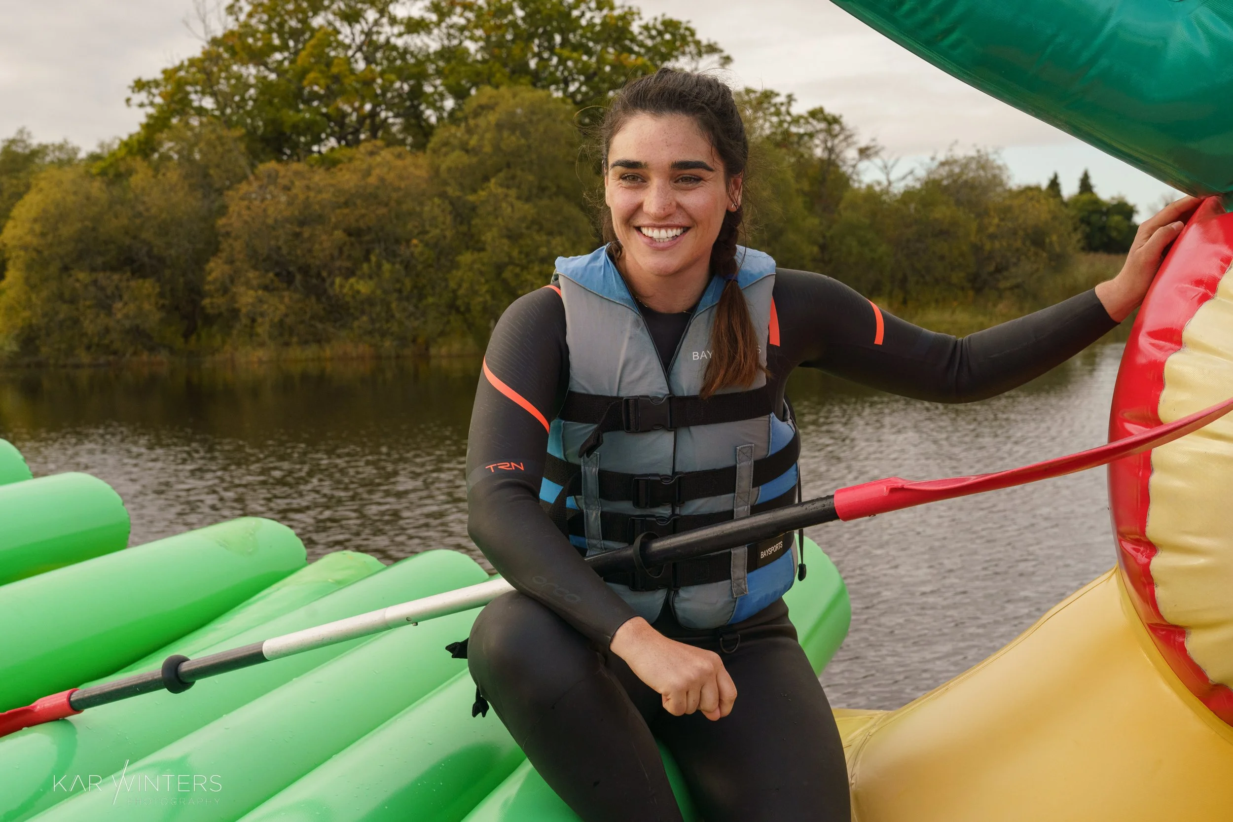 Woman smiling on a green inflatable raft holding a paddle on a river with trees in the background.