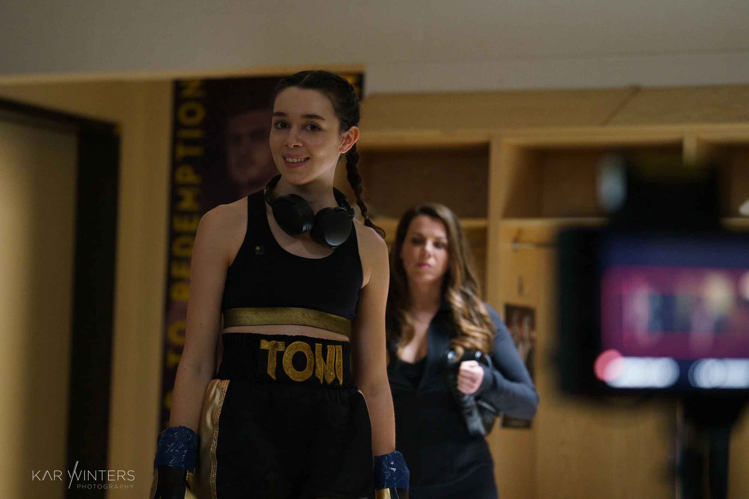 Young woman in boxing gear with black and gold shorts labeled 'TOM', black tank top, and boxing gloves, smiling at camera. Another woman in casual clothing holding a camera stands in the background. The setting appears to be a gym or training area.
