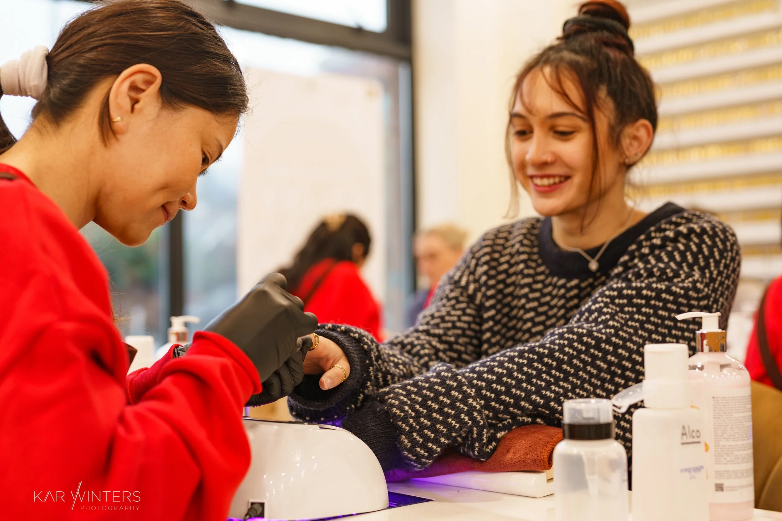 A young woman receives a manicure from a nail technician in a salon. The woman is smiling, wearing a black and beige sweater, and the technician is wearing a red uniform and black gloves. Various bottles of nail products are on the table.