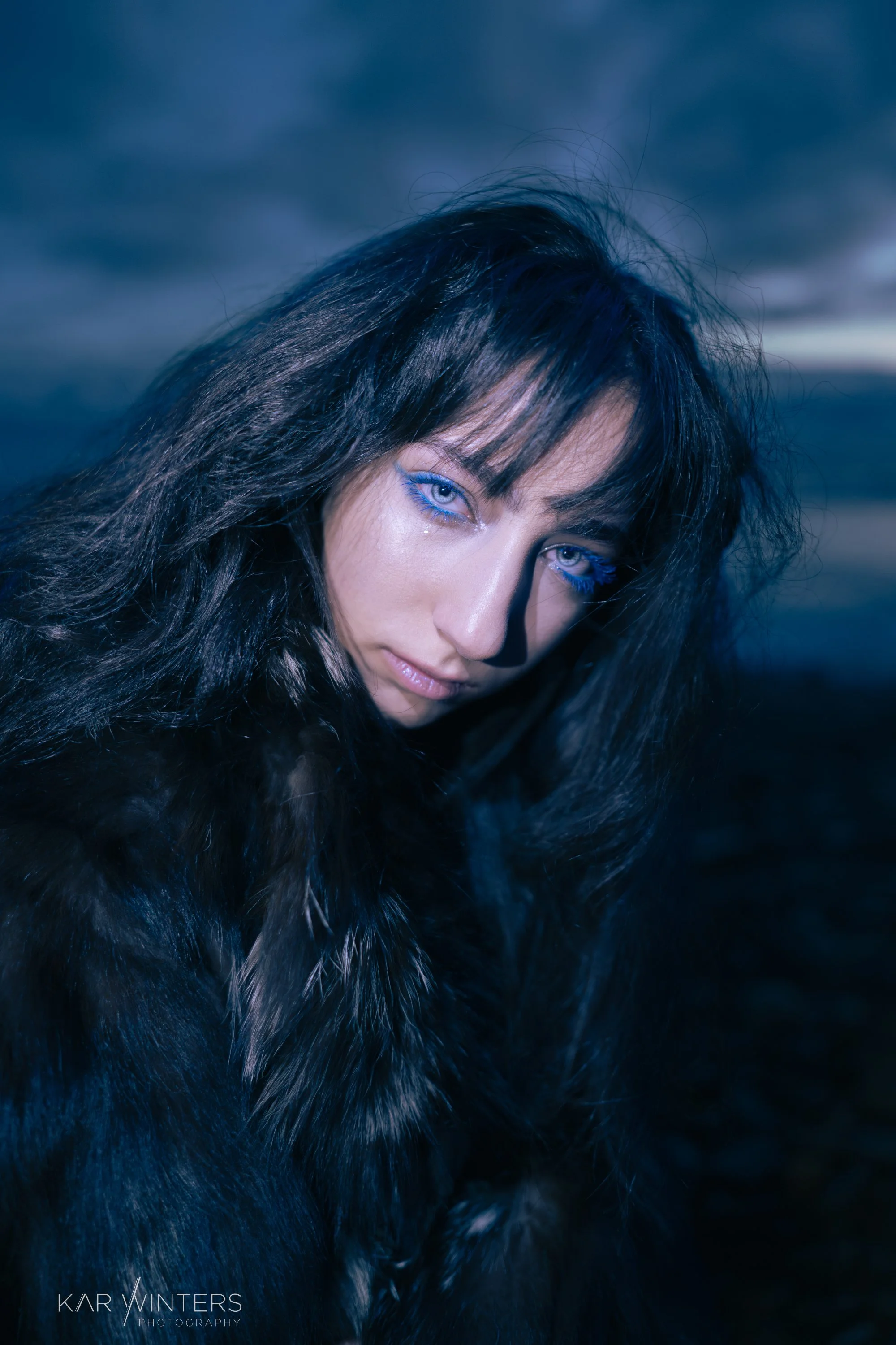 A woman with blue eyes and dark, messy hair, wearing a dark fur coat, outdoors during dusk or evening with cloudy sky.