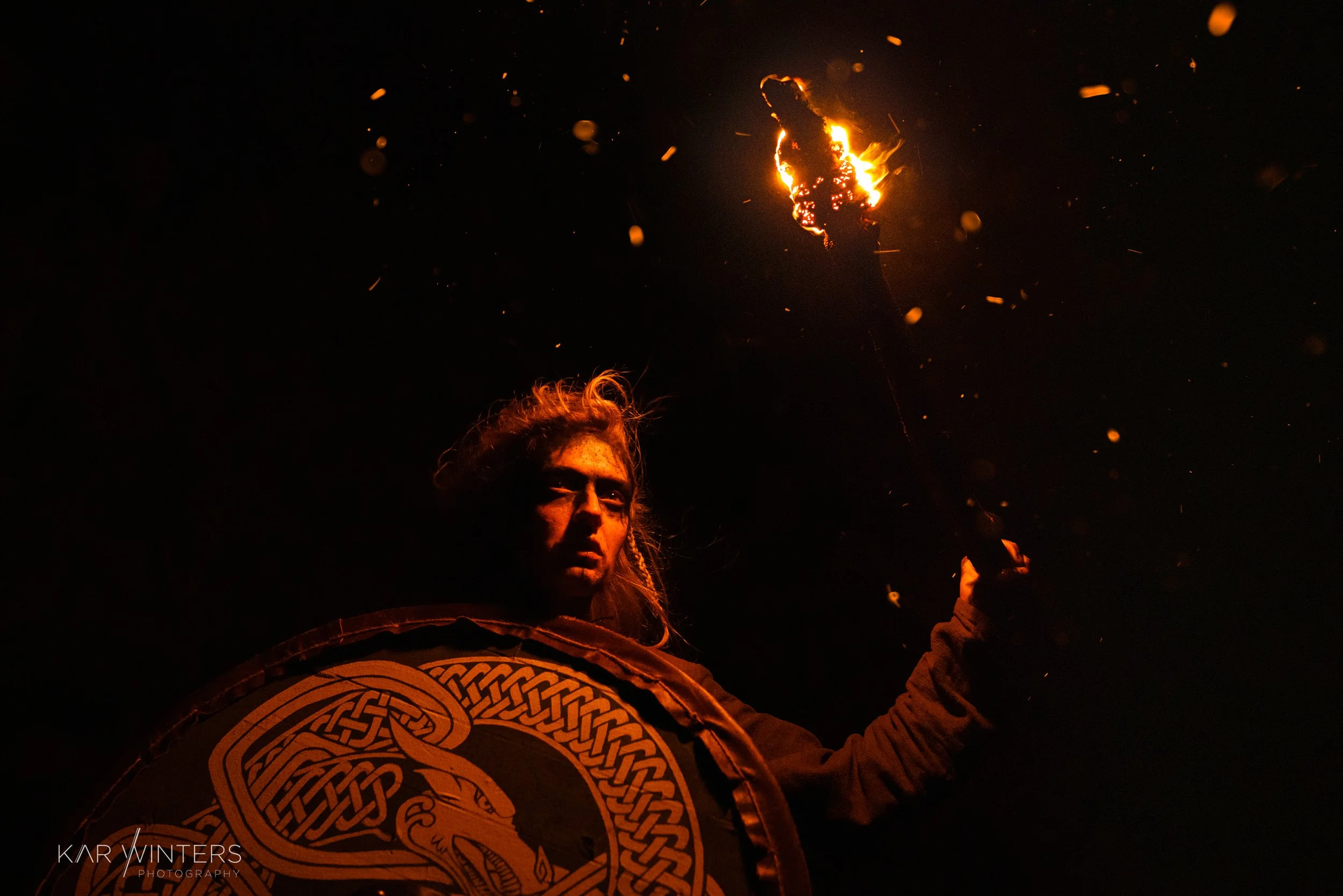A person holding a shield with a dragon design in front of a dark background, holding a flaming torch aloft. The scene is dimly lit, highlighting the fire and the person's intense expression.