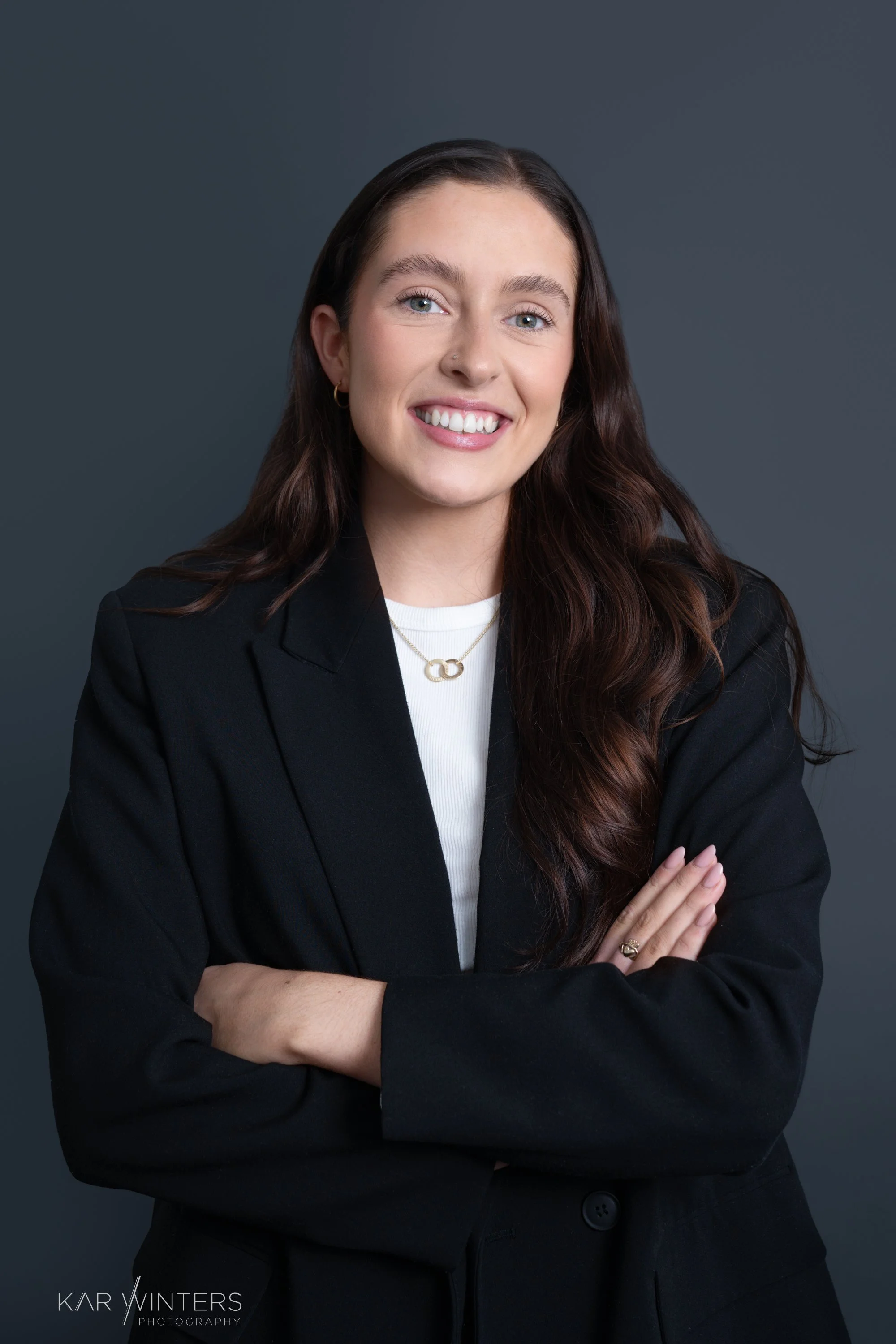 A woman with long brown hair smiling, wearing a black blazer, white shirt, and gold jewelry, crossing her arms against a dark gray background.
