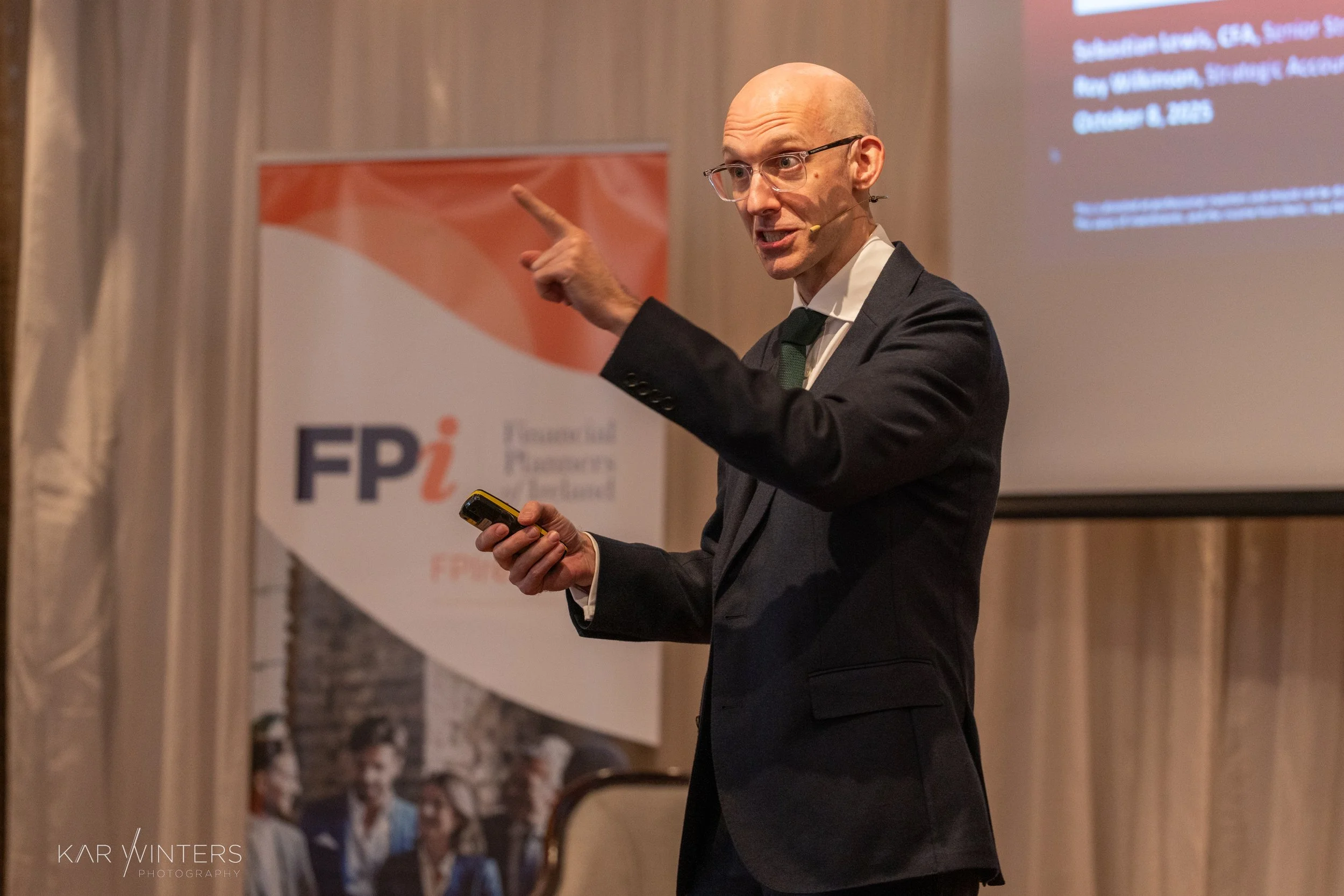 A man in a suit giving a presentation at a conference, pointing upwards with his left hand and holding a remote control in his right hand, with a screen and a FPi banner in the background.