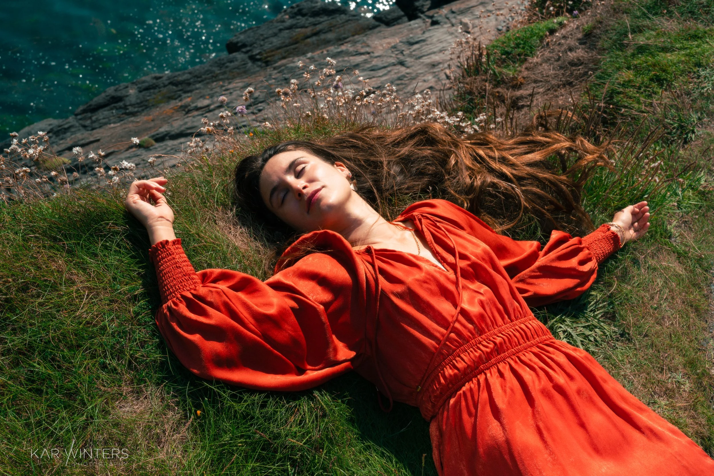 A young woman with long brown hair lies on her back on a grassy patch near a rocky shoreline, wearing an orange dress with puffy sleeves and a drawstring waist, with her eyes closed and arms relaxed.