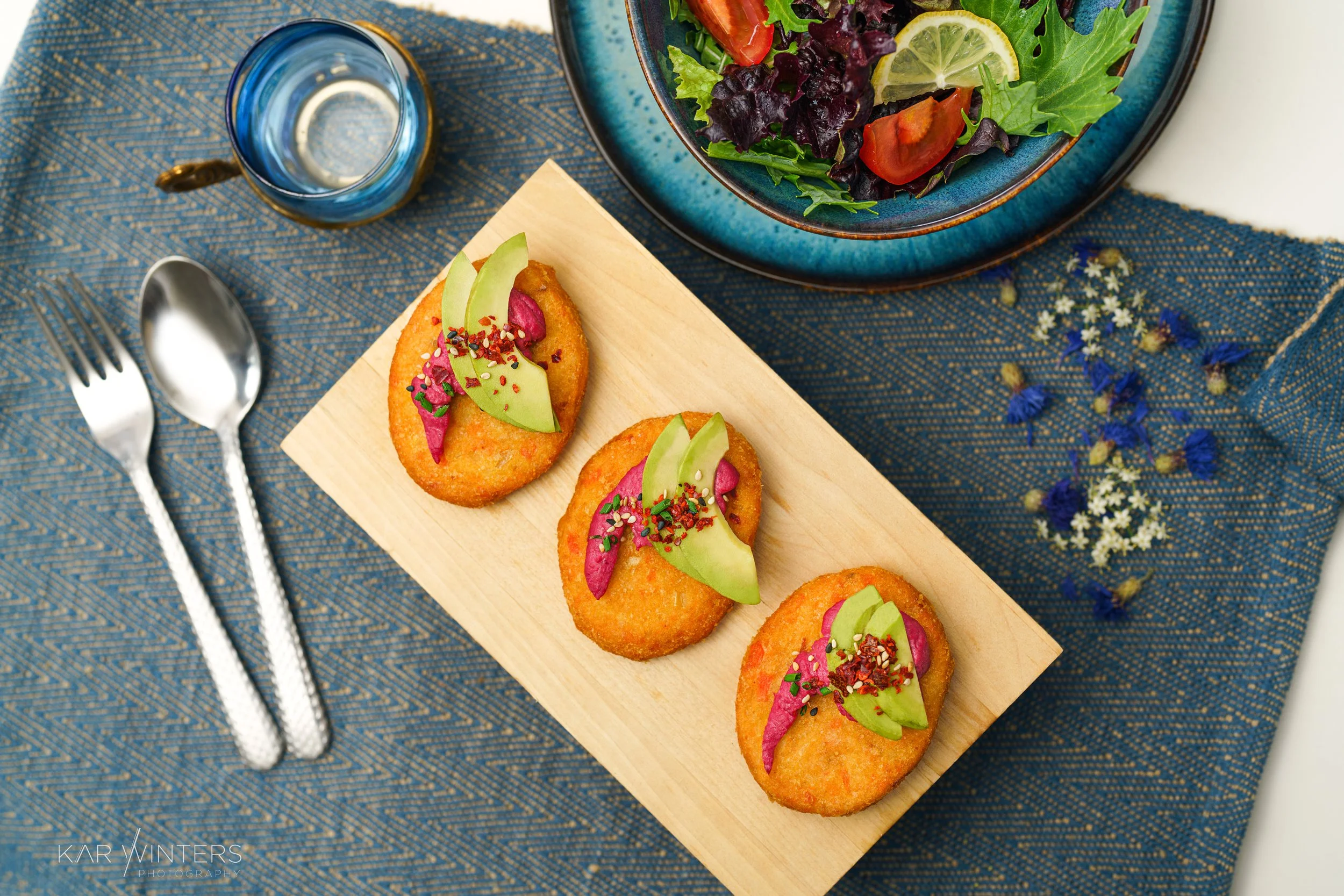 Three pieces of breaded fried food topped with avocado slices, pink pickled vegetables, and sprinkled with seeds, served on a wooden board. There is a blue patterned cloth underneath, a fork and spoon on the left, a glass of water, and a bowl of mixe