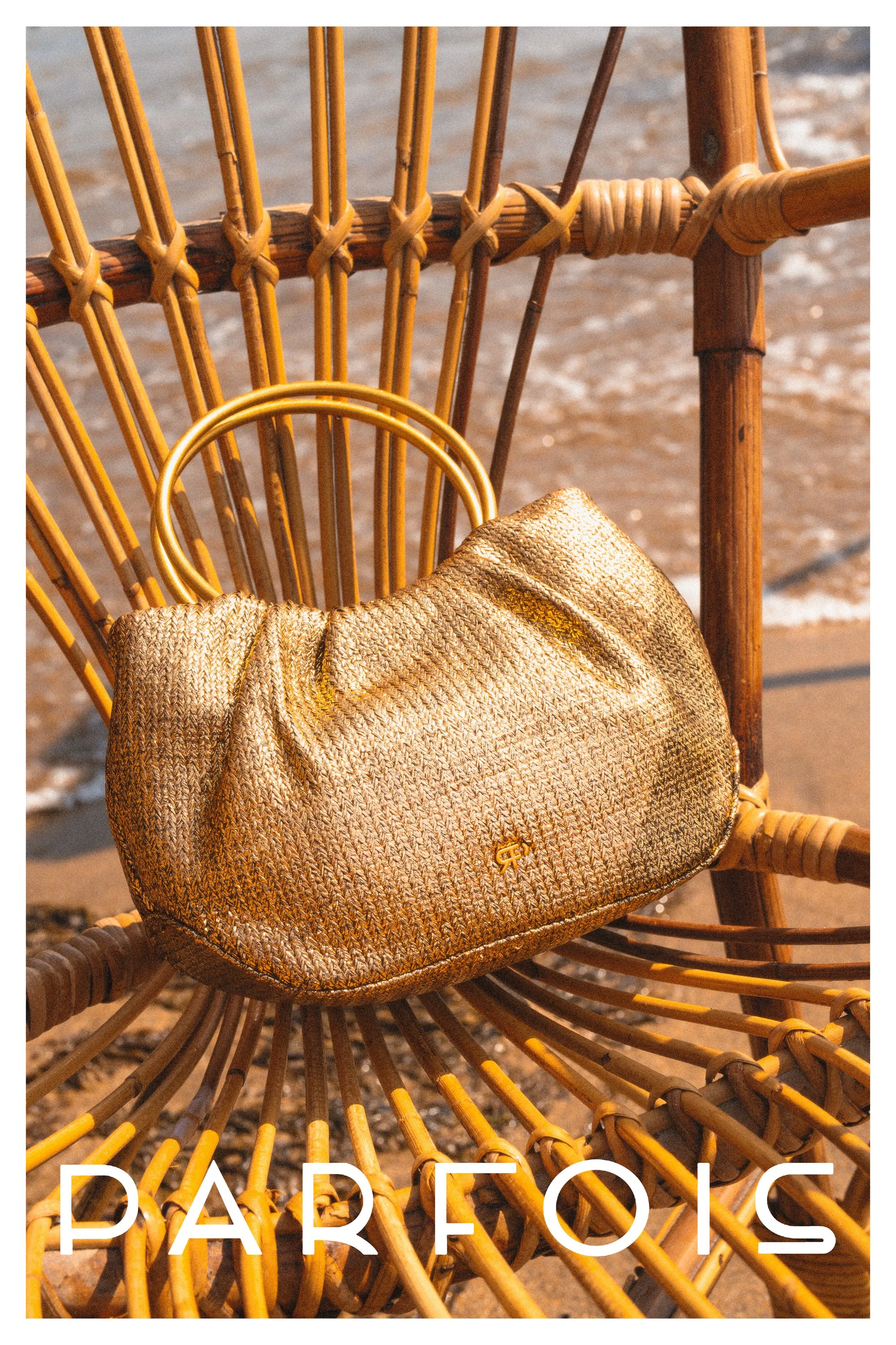 A woven beige and gold Balmain handbag resting on a wicker beach chair near the ocean.