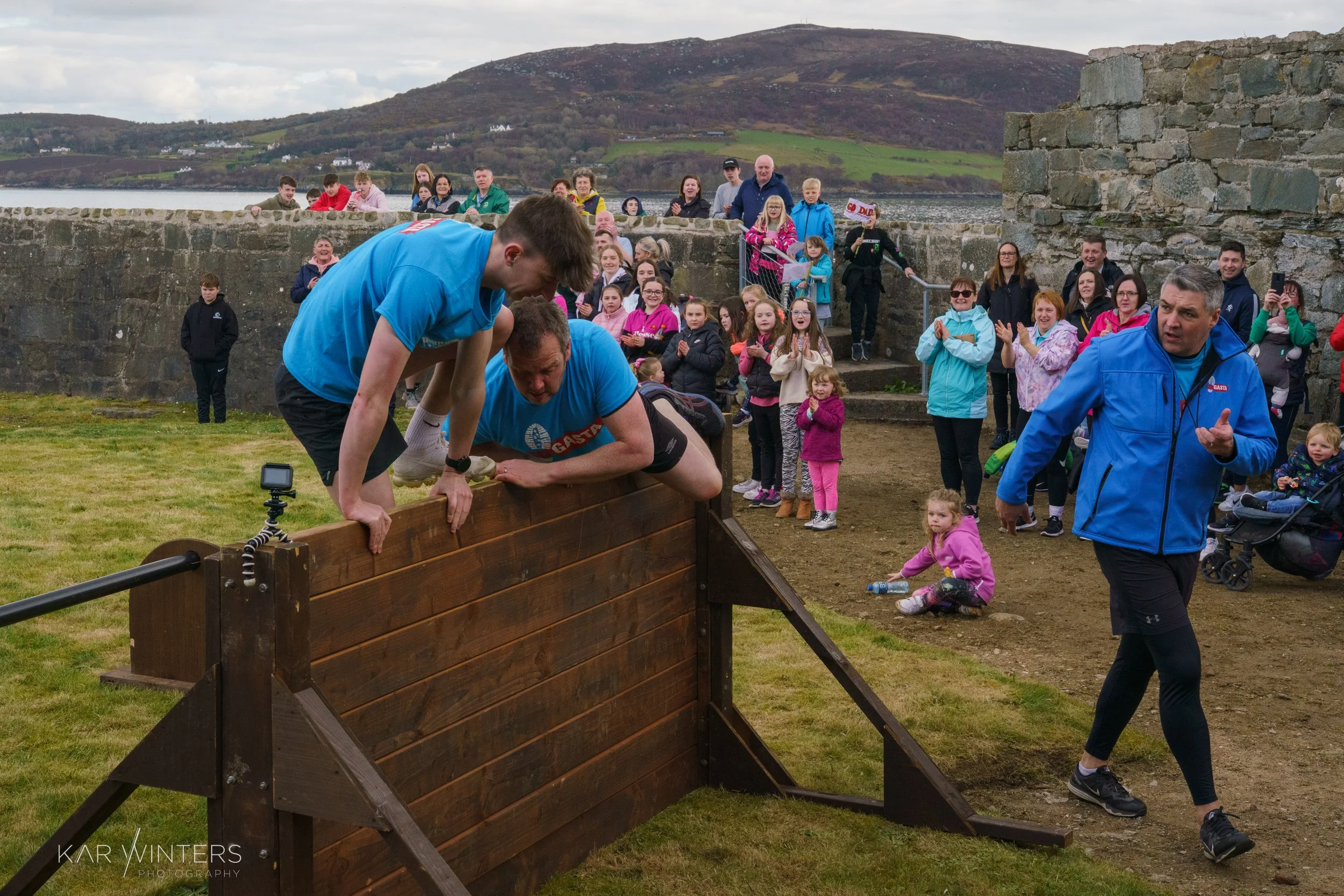 Two men in athletic clothing help each other over a wooden wall obstacle during a race, with a large crowd of spectators watching behind them on a grassy outdoor area, with hills and water in the background.