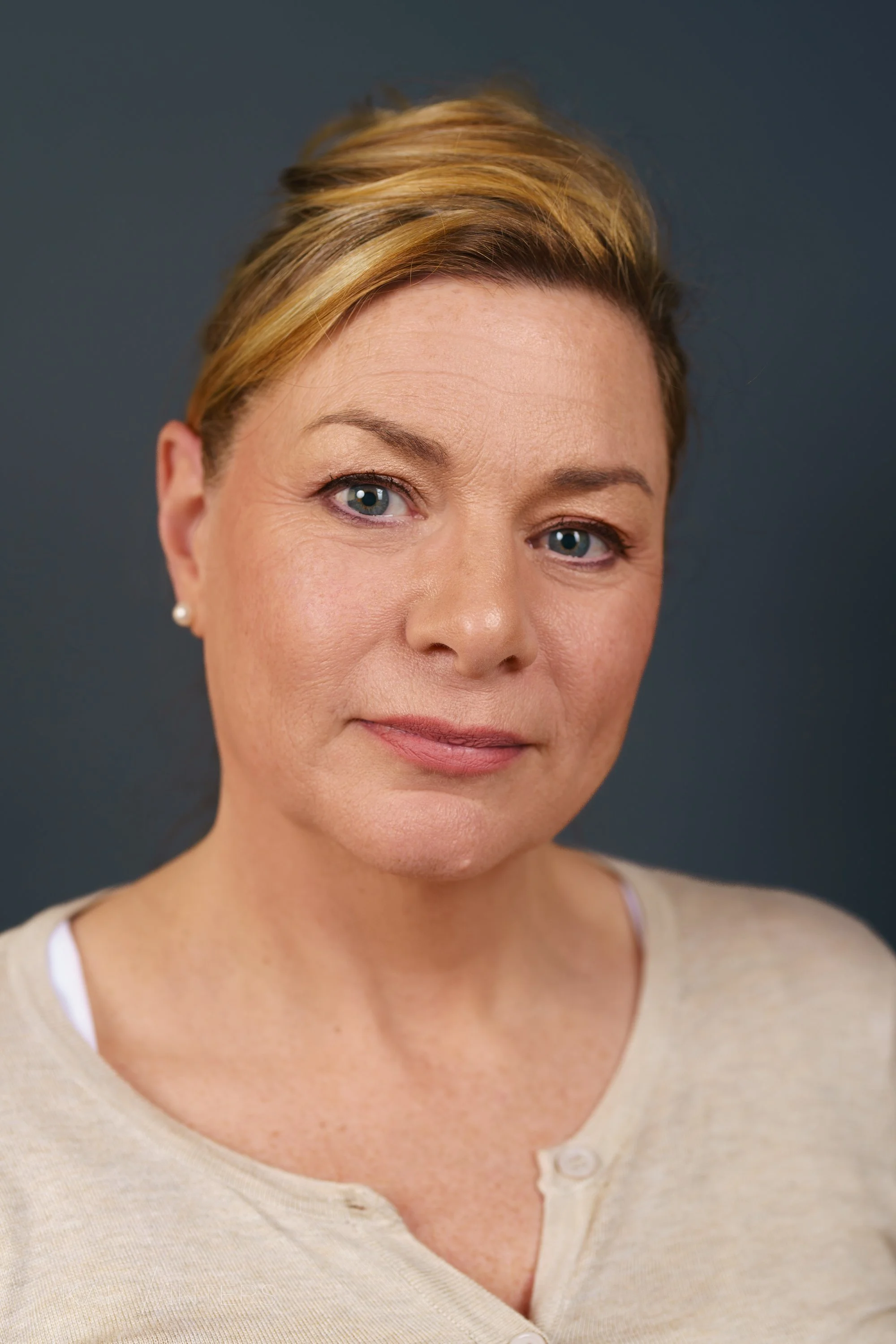 Close-up of a middle-aged woman with short blonde hair, blue eyes, and light skin, wearing a beige button-up sweater and pearl earrings, against a dark background.