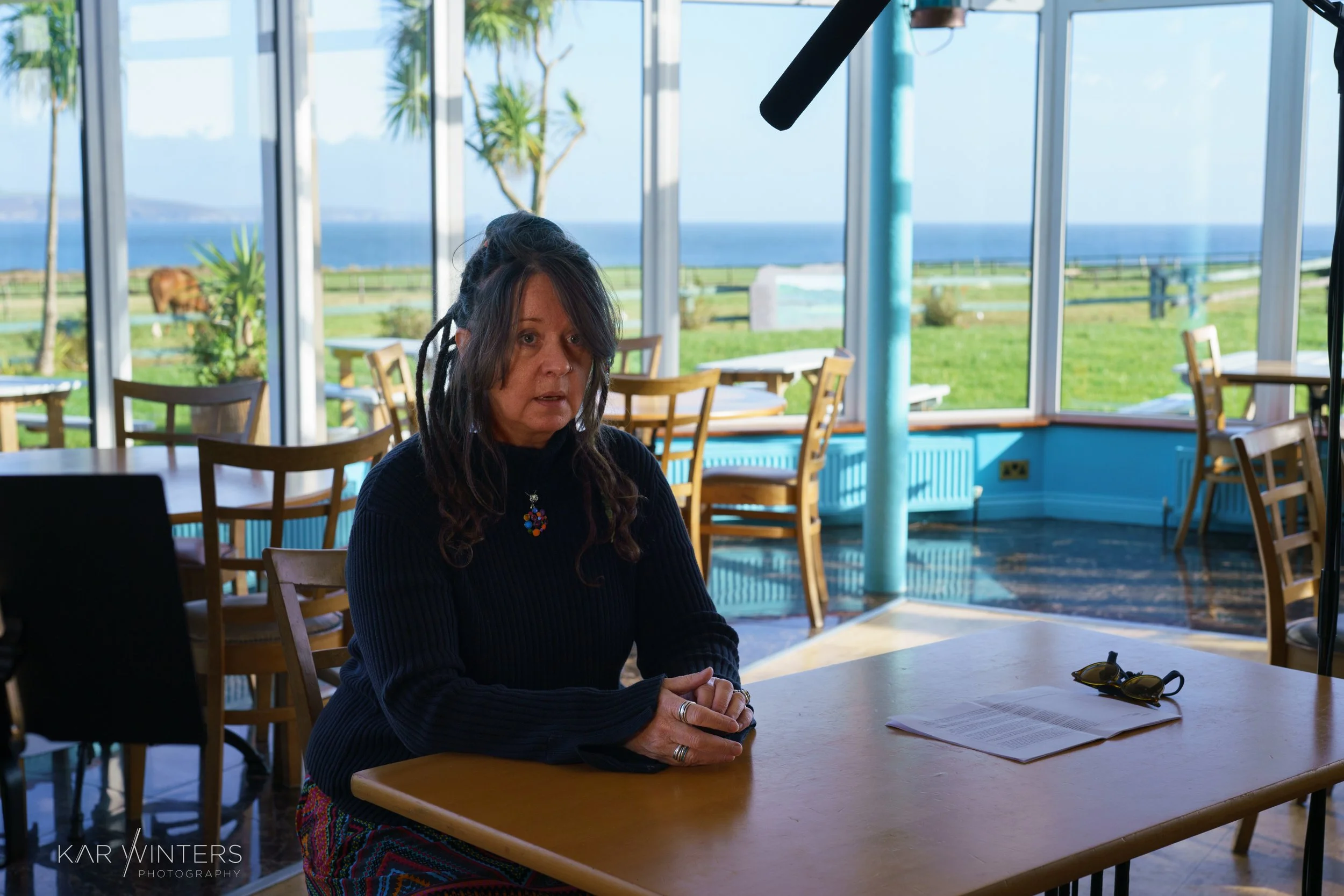 A woman with long, dark dreadlocks and wearing a dark sweater sits at a table in a restaurant or cafe with large windows. Outside, there is a view of a green lawn, a wooden fence, and the ocean in the distance. On the table, there is a sheet of paper