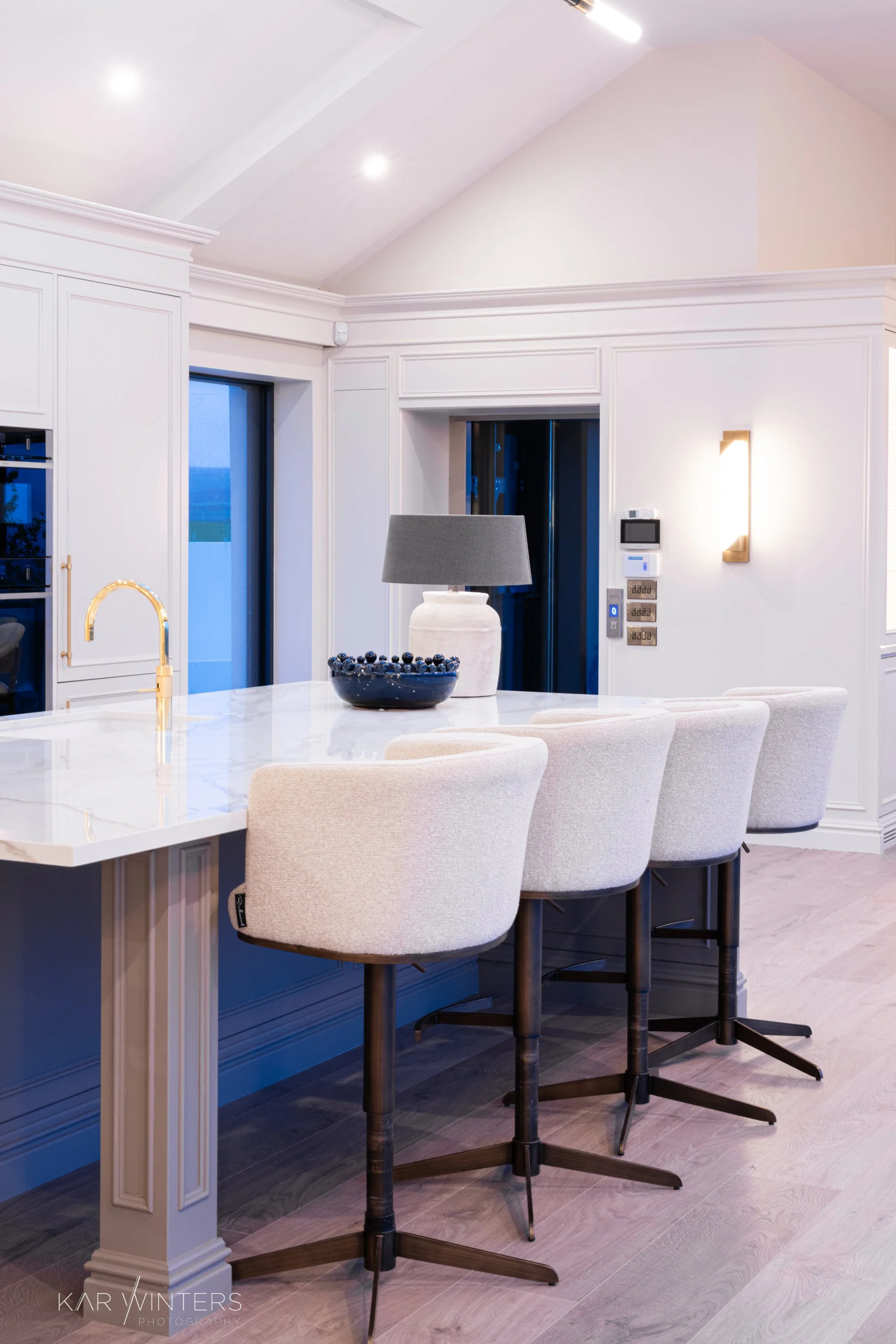 Modern kitchen with white cabinetry, marble countertop island, four beige bar stools, a black bowl with blueberries, a white vase with a gray lamp, and a wall-mounted light fixture.