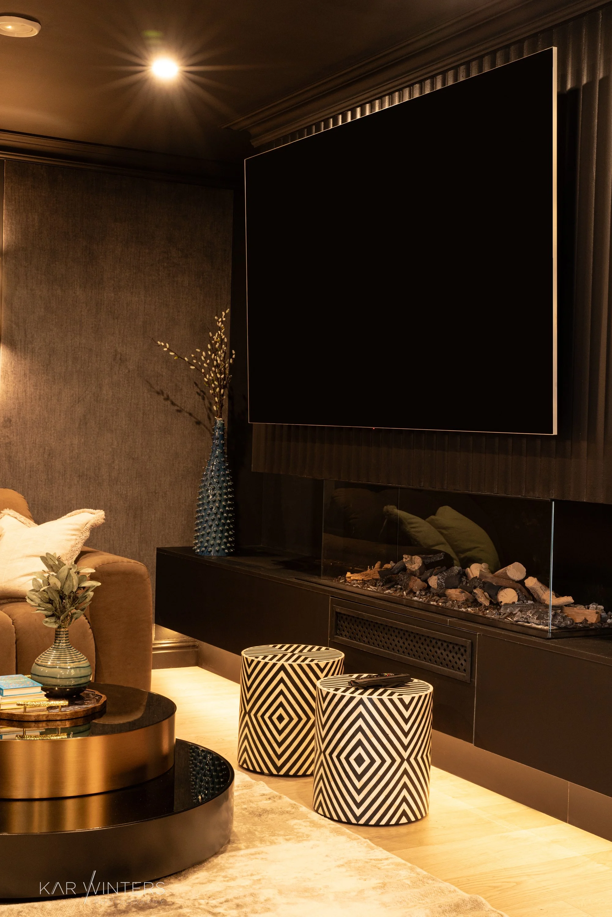 A modern living room with a black fireplace, large wall-mounted TV, a brown sofa with pillows, a vase with decorative branches, a round coffee table, and two black and white patterned stools.