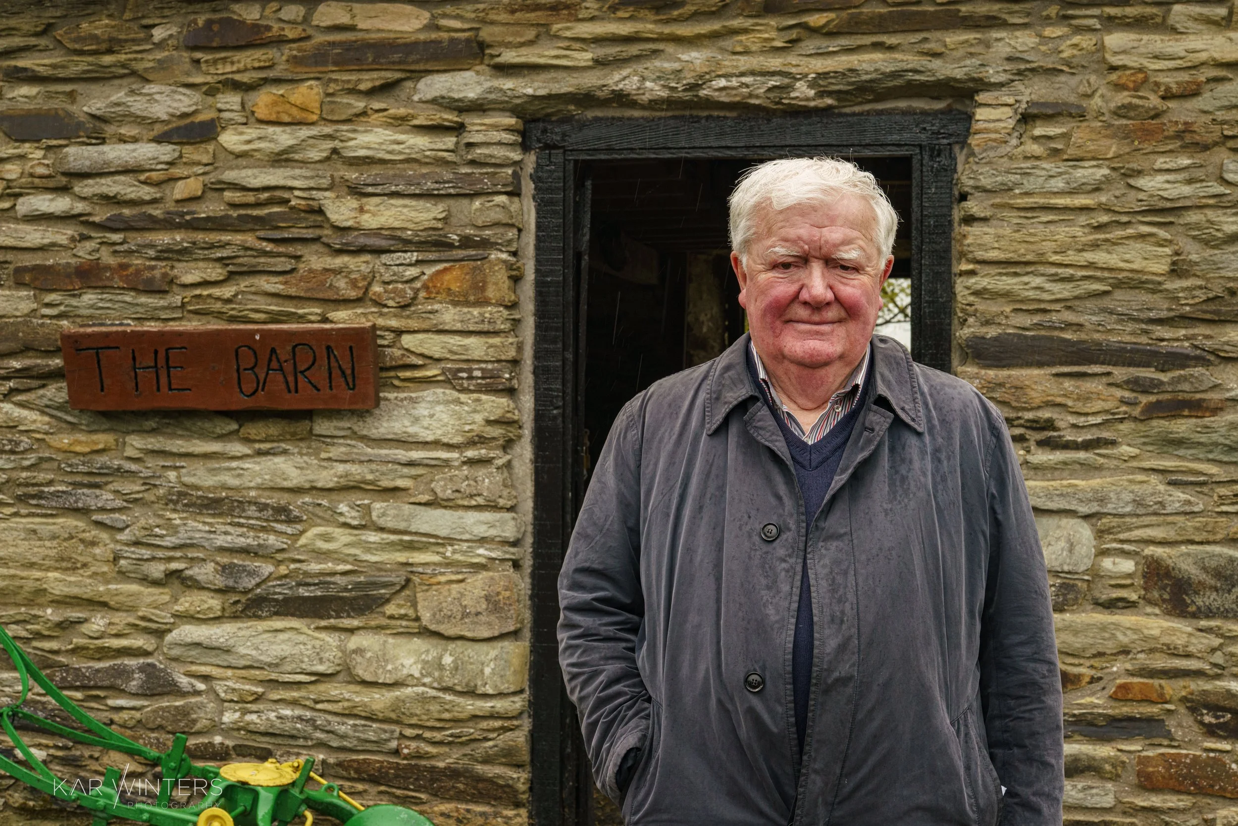 An elderly man with white hair wearing a gray jacket standing in front of a stone barn with a sign that reads 'The Barn' on the wall.