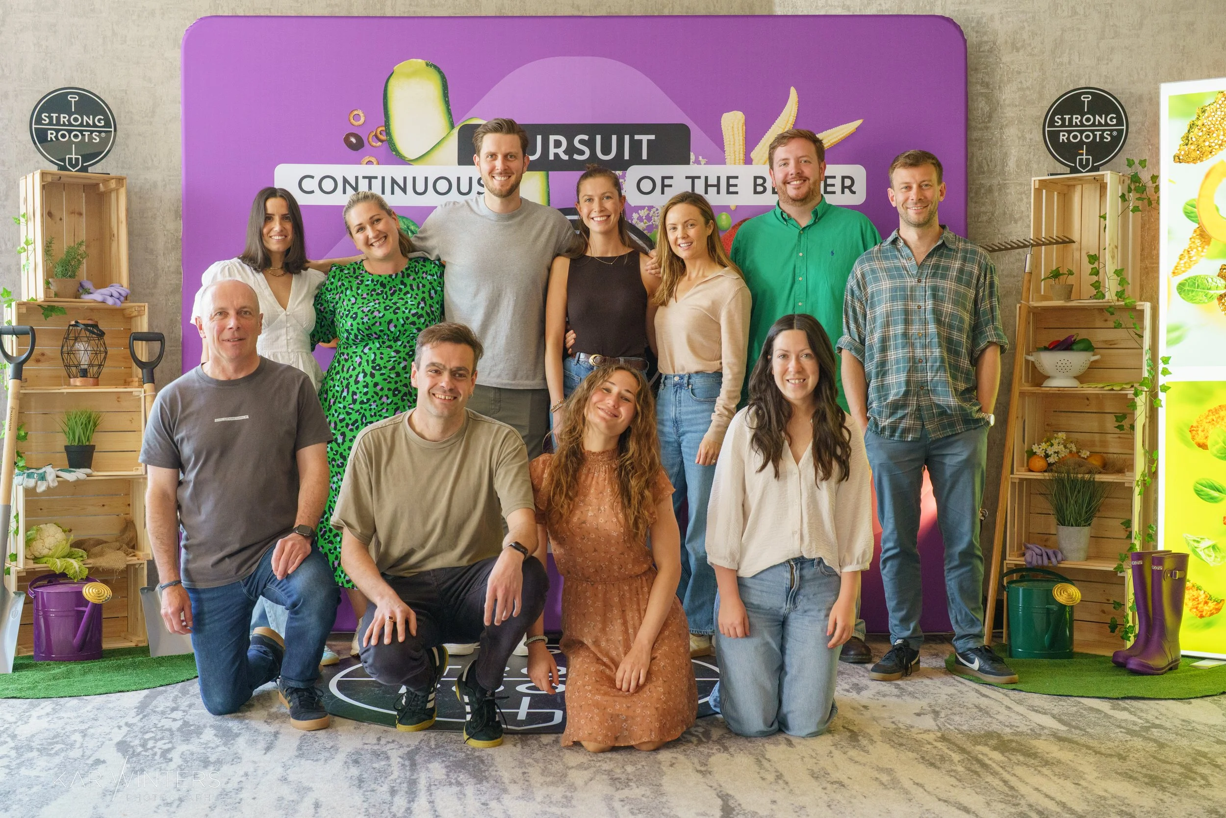 A group of fifteen people posing for a photo indoors, with a purple backdrop that has food-themed graphics and the words 'Pursuit of the Better' and 'Continuous'. They are standing and sitting in front of shelves with plants and gardening tools.