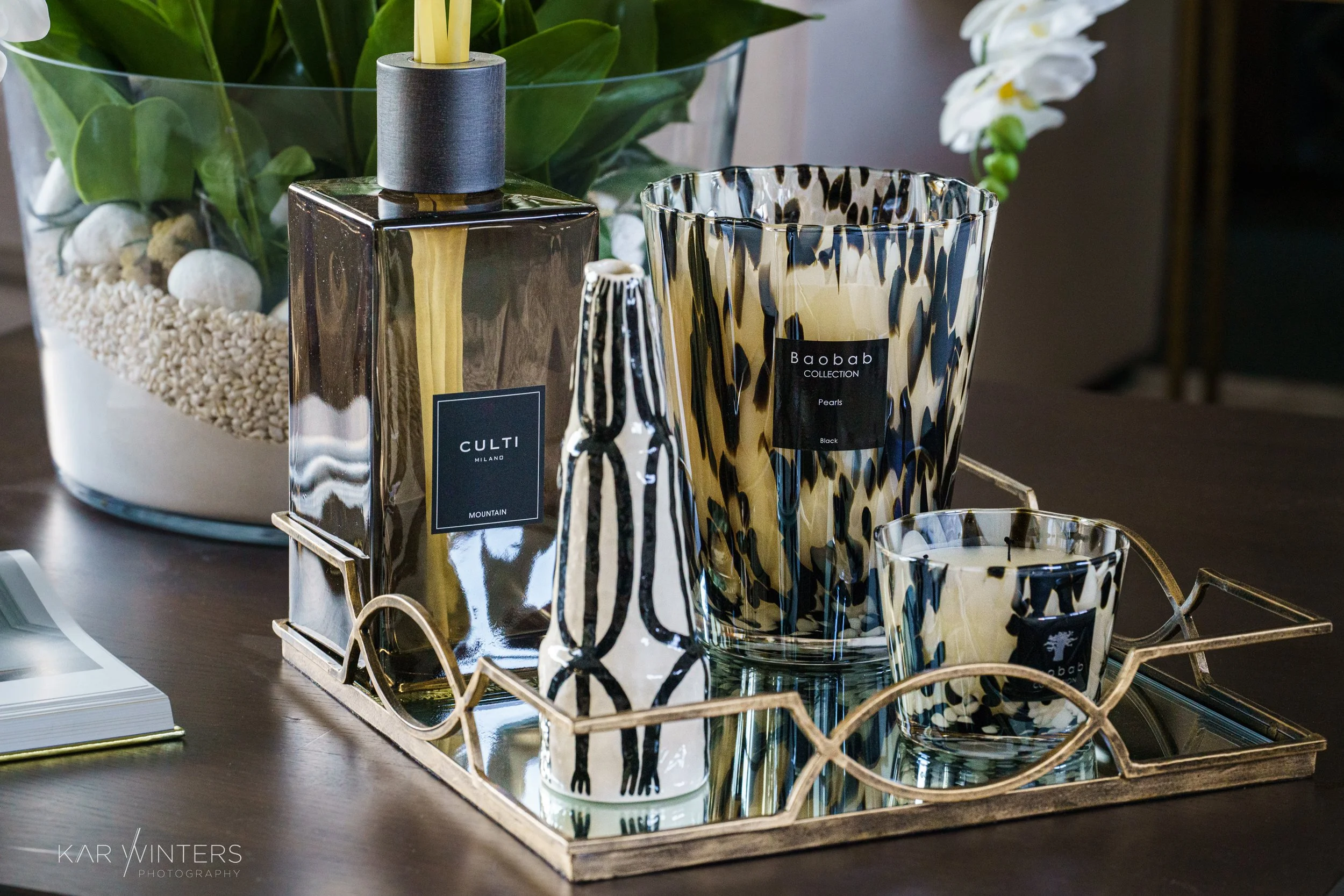 Decorative glass vases and candle holders with black, white, and beige abstract patterns are arranged on a gold tray on a dark wooden table, with a large clear glass bowl containing plants and white stones in the background.