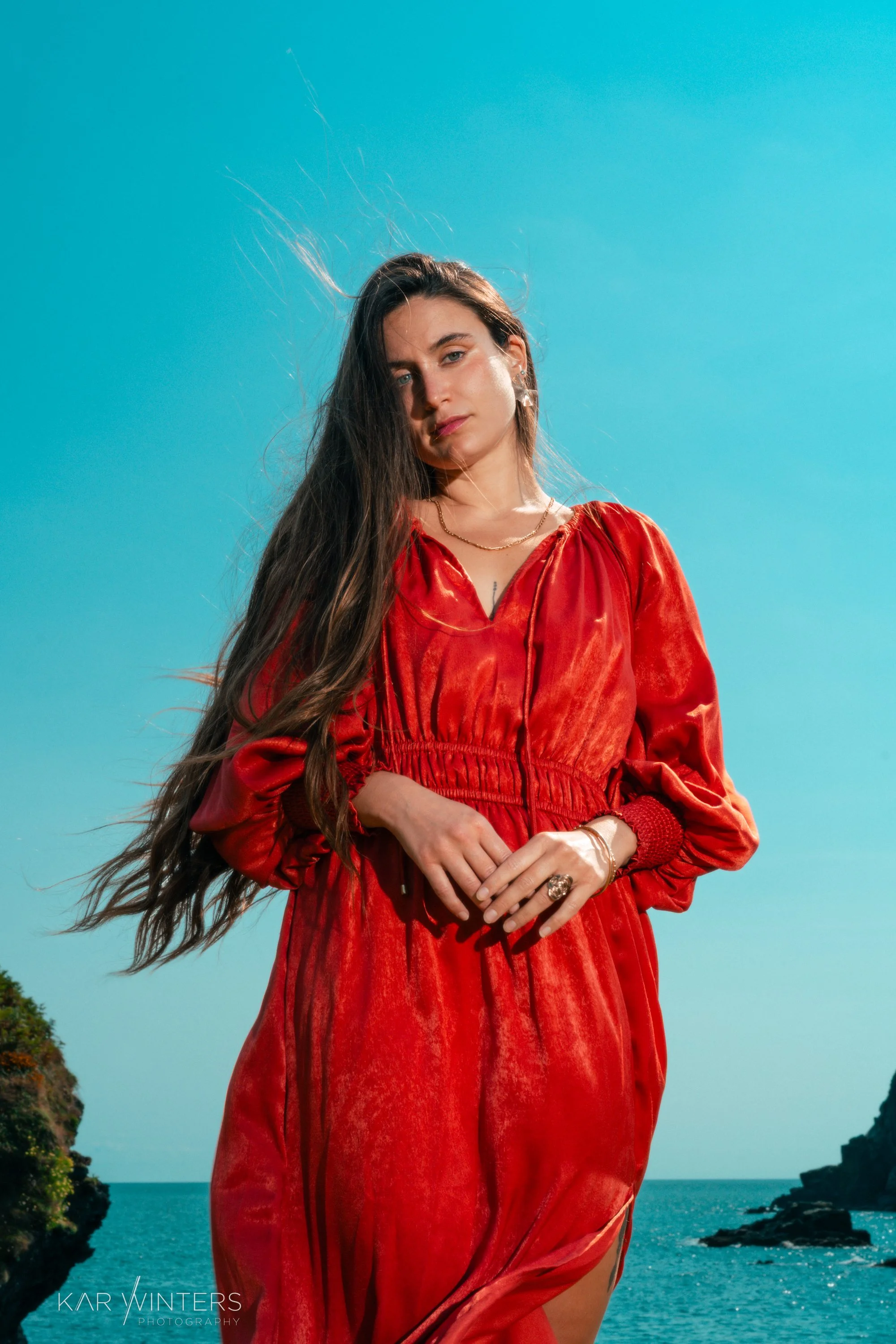 A woman in a red dress standing near the ocean with cliffs and blue sky in the background.