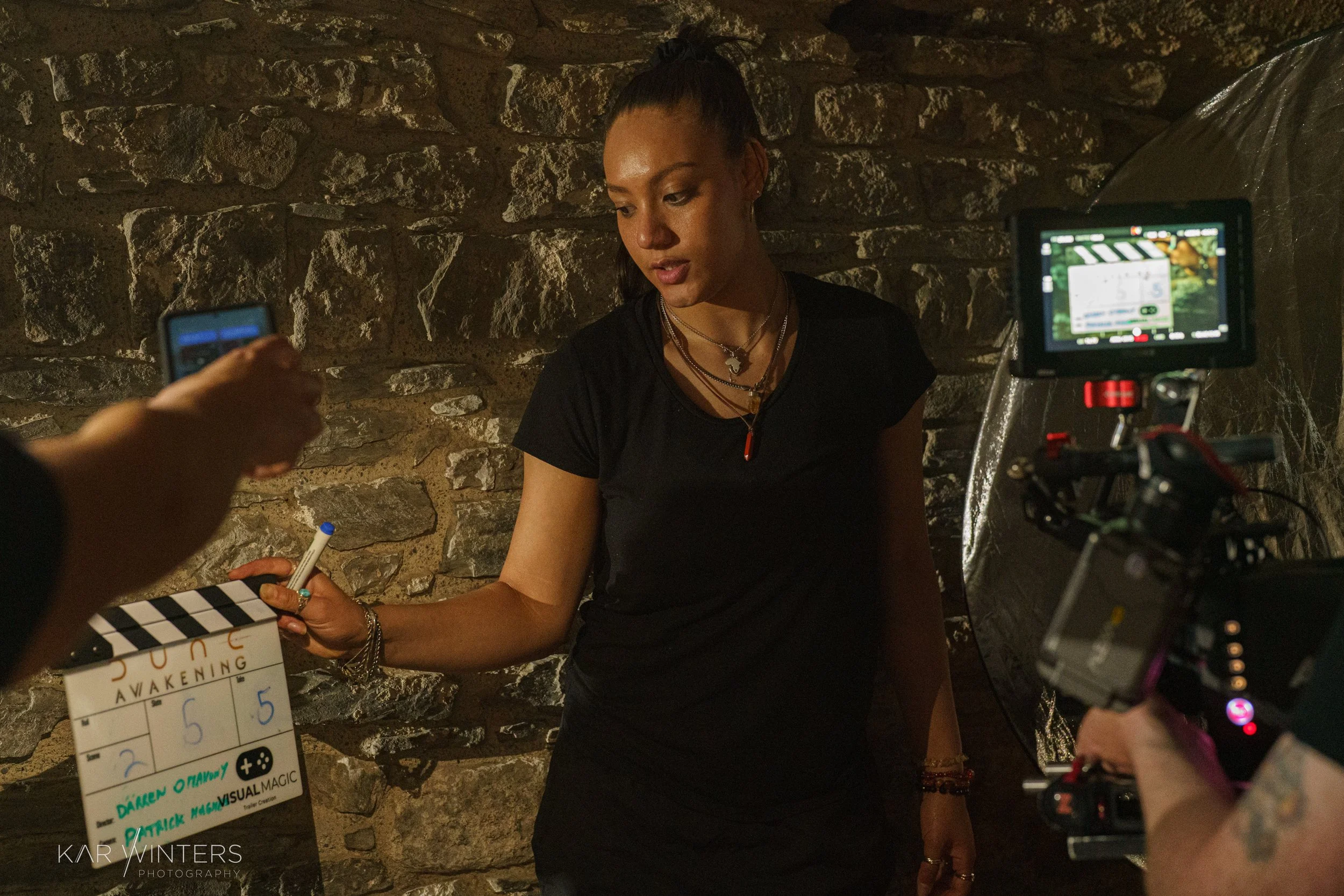 A woman is being photographed or filmed against a stone wall, holding a marker and a small slate with writing on it. She is wearing a black shirt, multiple necklaces, and bracelets, and has dark hair tied back. A person with a tattooed arm is operati