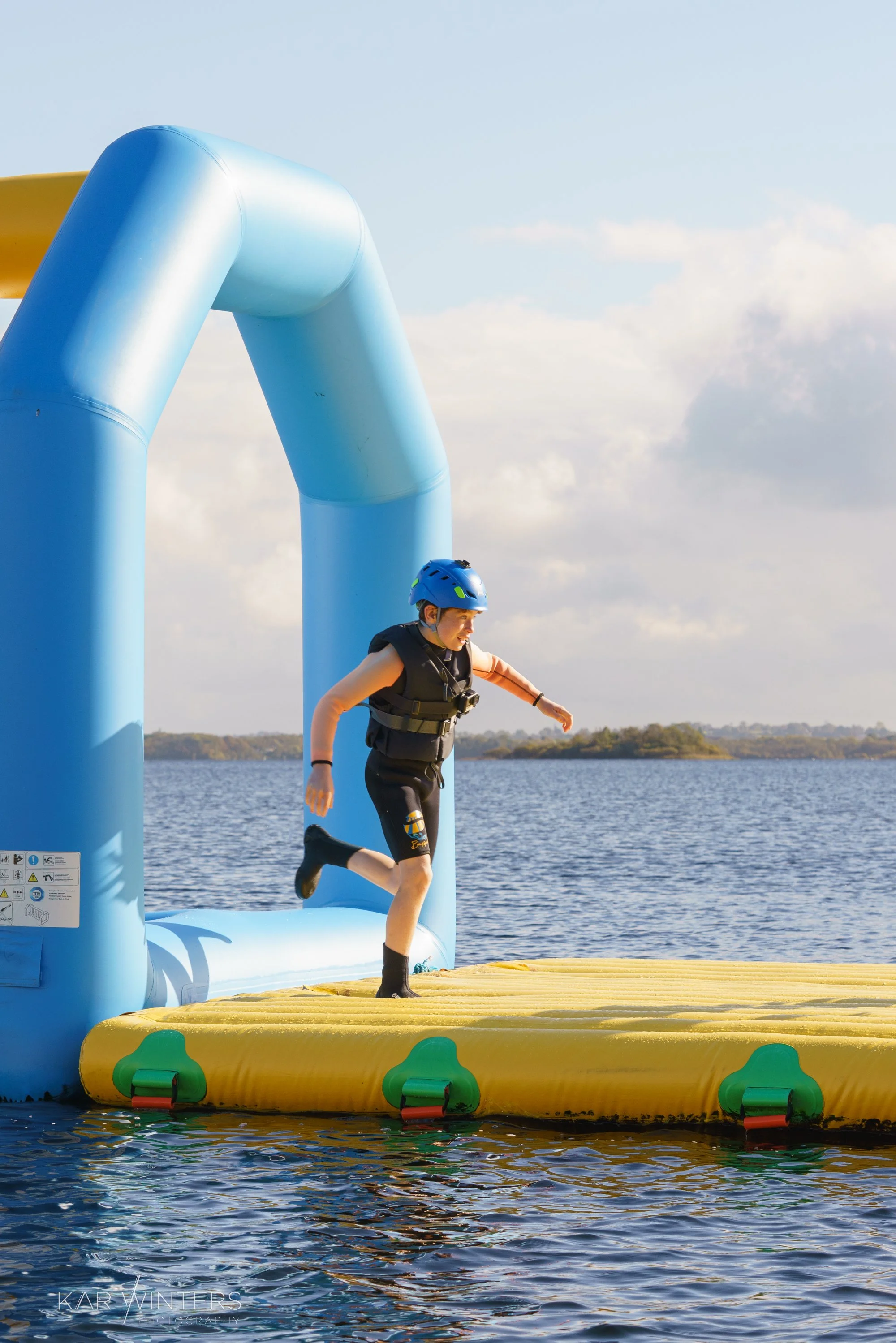 Young girl in a blue helmet and life jacket running on an inflatable water trampoline at a lake.